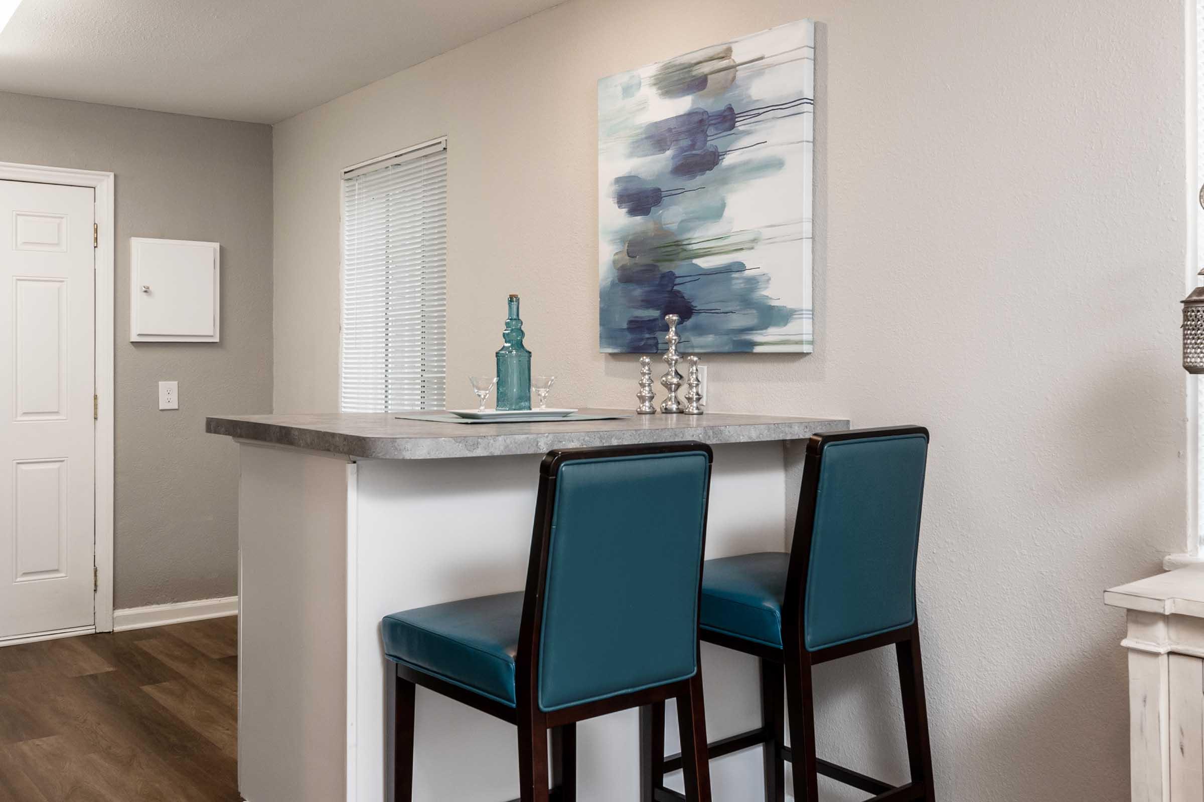 A modern kitchen area featuring a bar counter with two teal chairs. Above the counter, there is an abstract painting with blue and gray hues. The wall is painted a light color, and a window with blinds is visible. The space has a clean, stylish appearance, suitable for entertaining or casual dining.