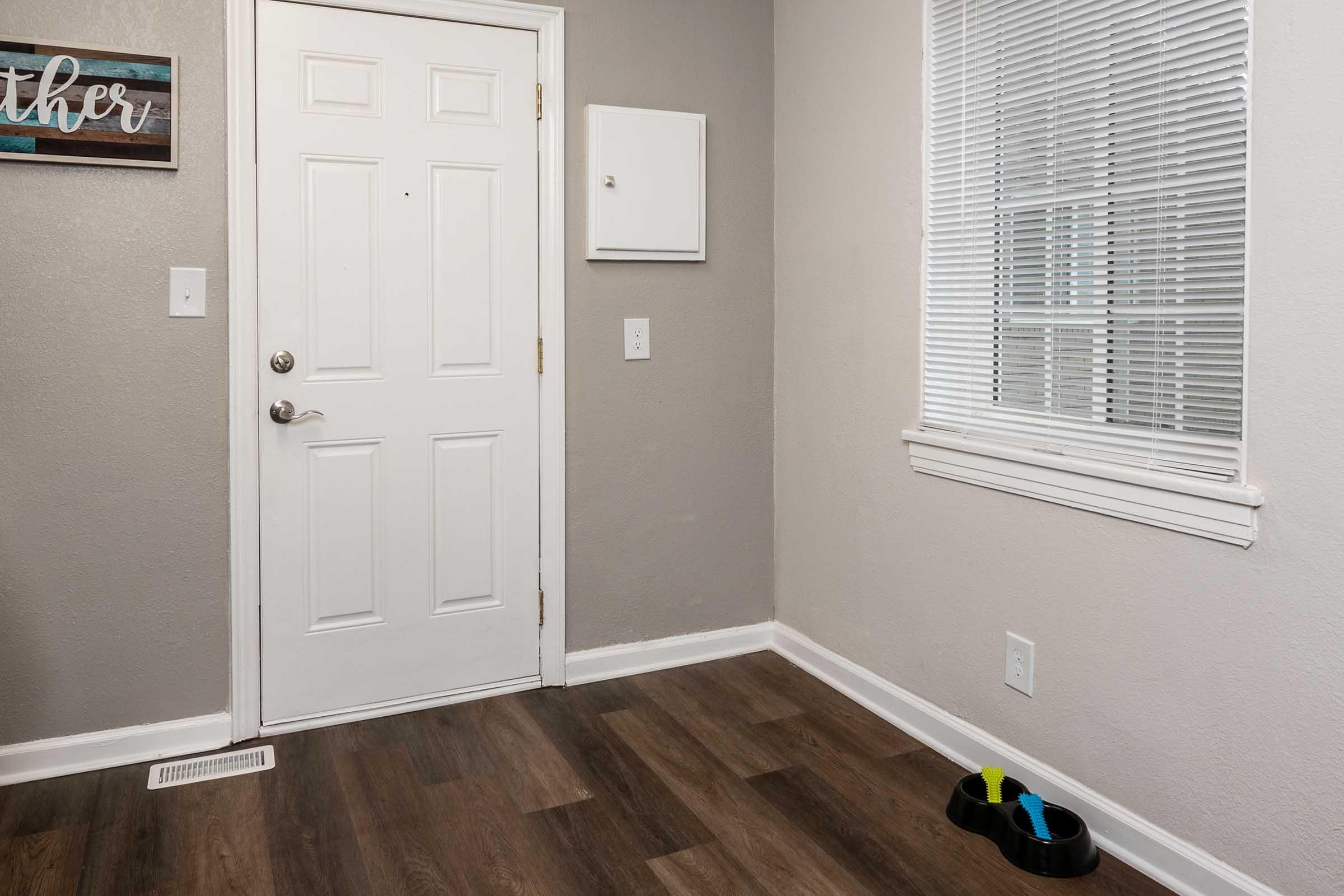 A small entryway featuring a white door with a silver handle, a wall-mounted mail holder, and a window with blinds. The floor is made of wood laminate, and there are two pet bowls on the ground in the corner, colored yellow and green. The walls are painted a light gray.