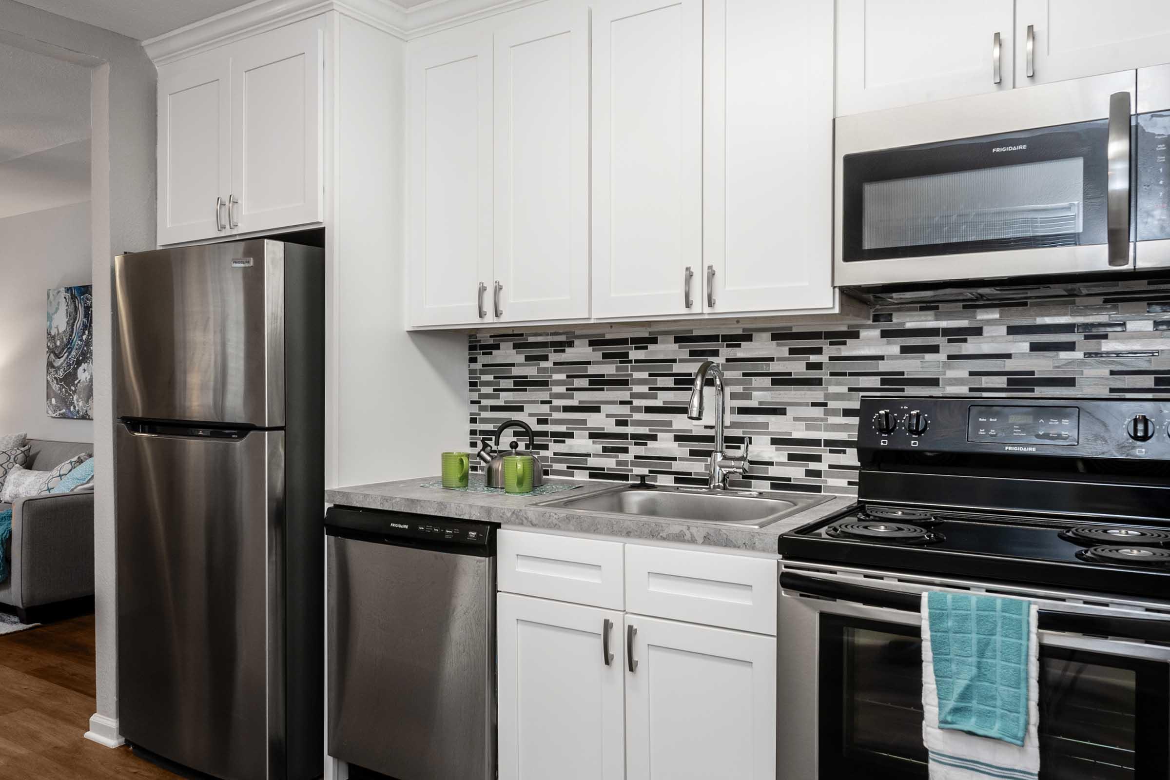 Modern kitchen featuring stainless steel appliances, including a refrigerator, dishwasher, and oven. The cabinetry is white, complemented by a gray and black mosaic tile backsplash. A light blue towel hangs from the oven, adding a pop of color. The space is well-lit and designed for functionality.
