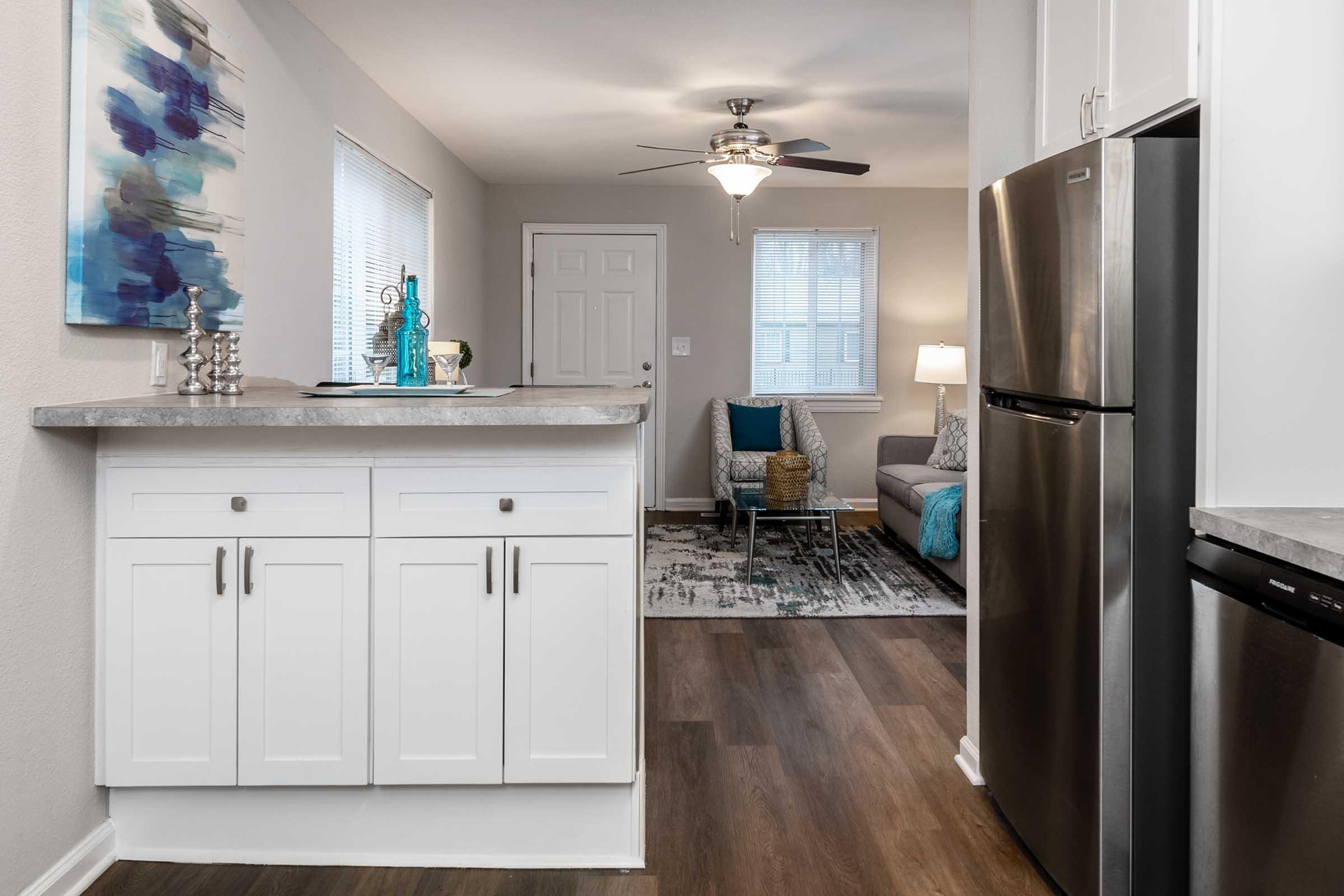 Modern kitchen view, featuring white cabinetry and a stainless steel refrigerator. A living area is seen in the background with a gray couch, decorative throw pillows, and a light fixture. Natural light comes through the windows, highlighting a contemporary aesthetic with blue accents.