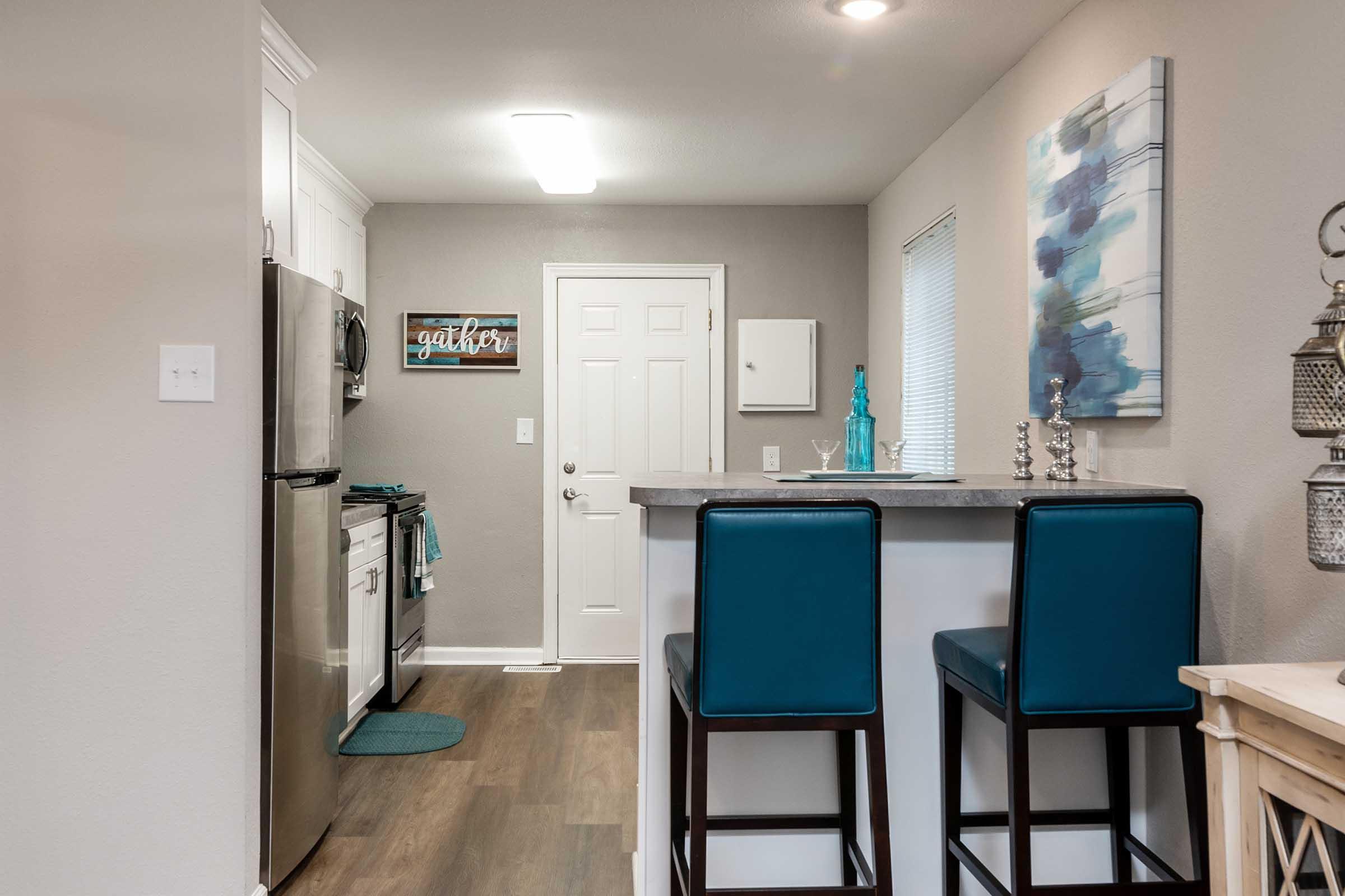 A modern kitchen with stainless steel appliances, including a refrigerator and stove. There's a small breakfast bar with two blue high-backed stools. The walls are painted light gray, and there's a decorative piece with blue accents. The entrance features a white door, and the space is well-lit and inviting.