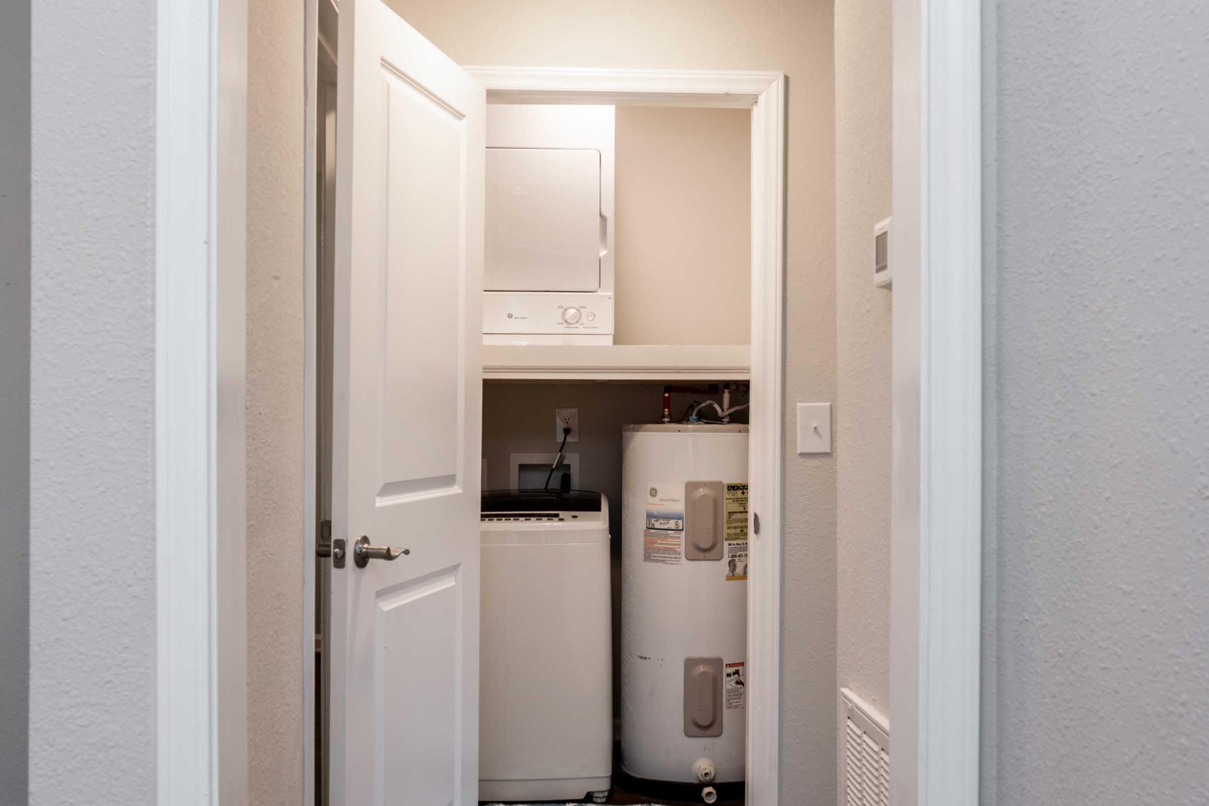 A small laundry room featuring a stacked washer and dryer unit on top and a water heater beside it. The room is accessed through a white door, with neutral-colored walls and trim, and a light source illuminating the space. There are visible electrical outlets and a vent in the room.