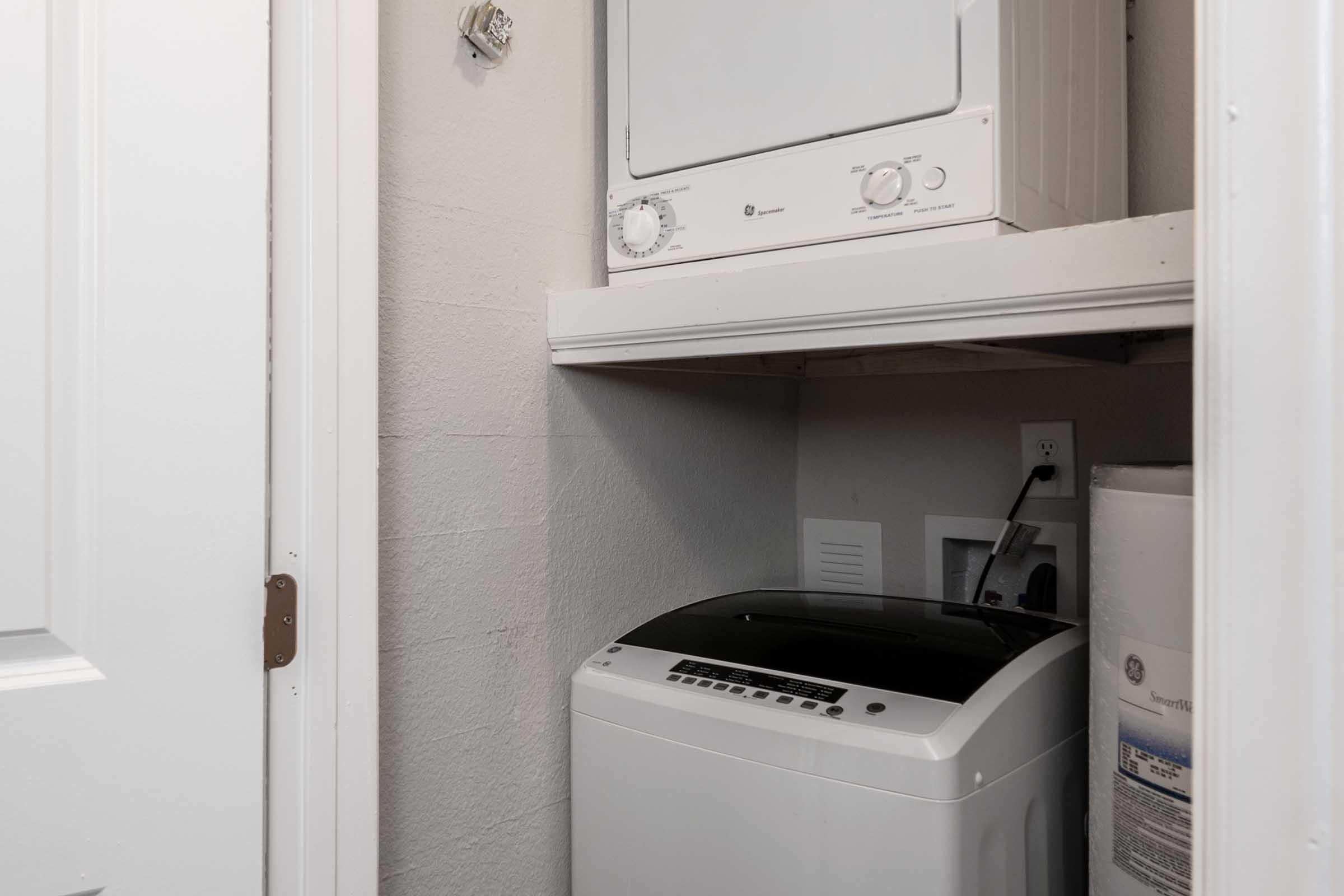 A small laundry area featuring a stacked washer and dryer on a shelf, with a top-loading washing machine below. A white water heater is visible to the right. The space is enclosed by a door, and there's an electrical outlet and venting on the wall. The walls are painted light gray.