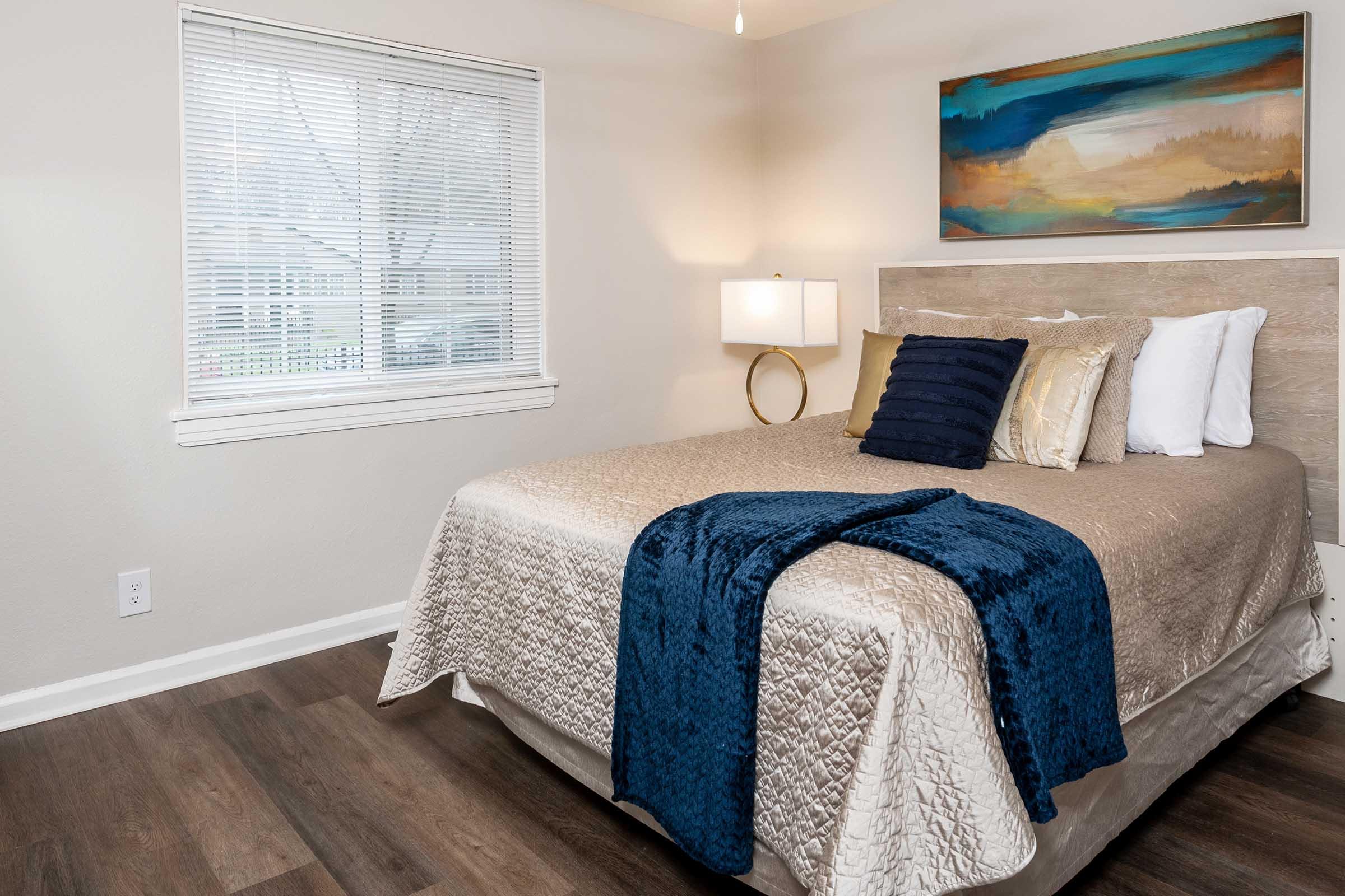A well-lit bedroom featuring a neatly made bed with a gold and cream quilt, blue throw, and decorative pillows. The room has a window with blinds, a modern lamp on a bedside table, and a colorful abstract painting on the wall above the bed. The flooring is wood laminate, adding warmth to the space.