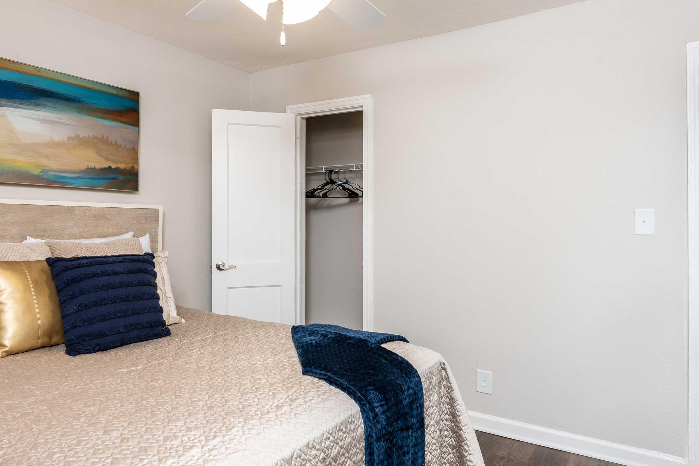 Cozy bedroom featuring a neatly made bed with decorative pillows and a soft blanket. A painting hangs above the bed, and a closet door is partially open, showing hangers inside. The walls are painted a light color, and the room has a ceiling fan for added comfort.