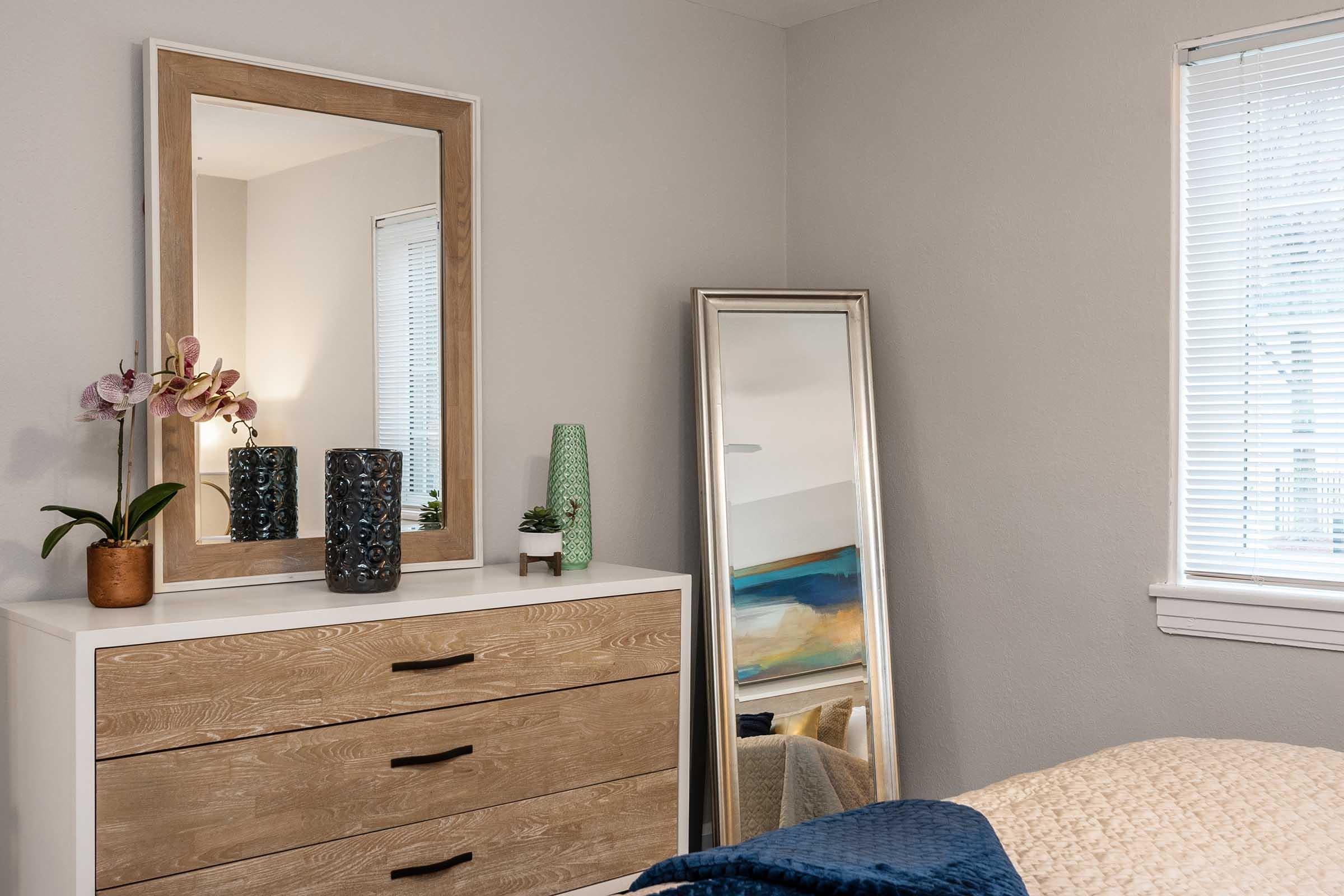 A cozy bedroom featuring a dresser with three drawers, a large mirror with a light wooden frame, a decorative black vase, a small potted plant, and an orchid. A tall, full-length mirror stands beside the dresser, while soft natural light filters through the window adorned with blinds. The room has a warm and inviting feel.