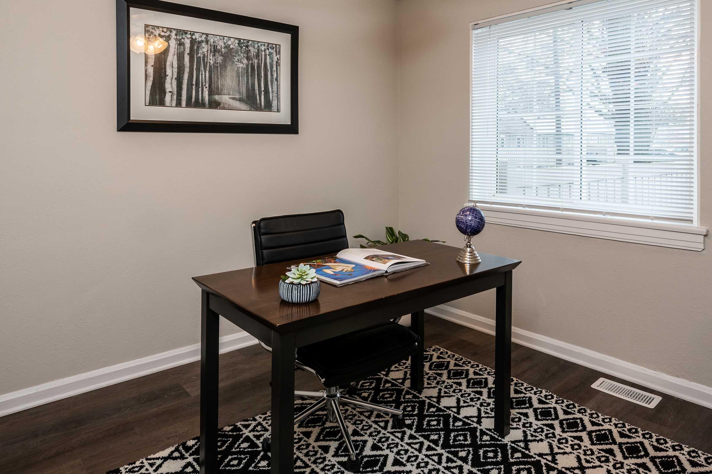 A modern minimalist home office featuring a sleek black chair and a wooden desk. On the desk, there is a decorative globe, a small plant, and an open book. A framed black-and-white photo of a forest hangs on the wall, and sunlight filters through a window with blinds, illuminating the space.