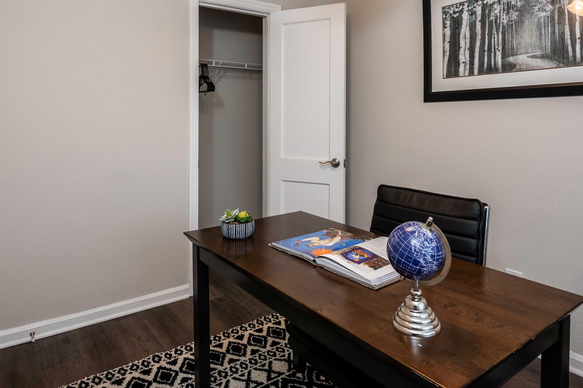 A modern home office space featuring a wooden desk with a decorative globe, a closed book with illustrations, and a small succulent plant. The room has a neutral color scheme with gray walls, wood flooring, and a framed black-and-white landscape picture. A closet door is visible, partially open, in the background.
