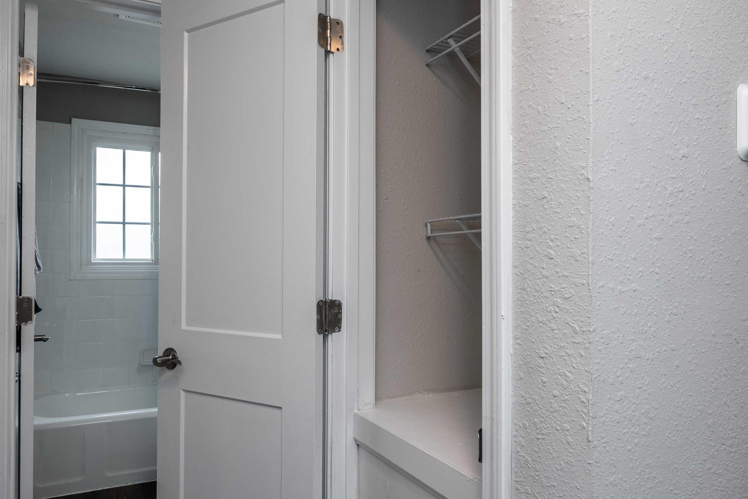 A partially open door reveals a bathroom with a bathtub and a window. Next to it is a small closet with wire shelving. The walls are painted in neutral tones, and the scene is well-lit, suggesting a clean and modern space.