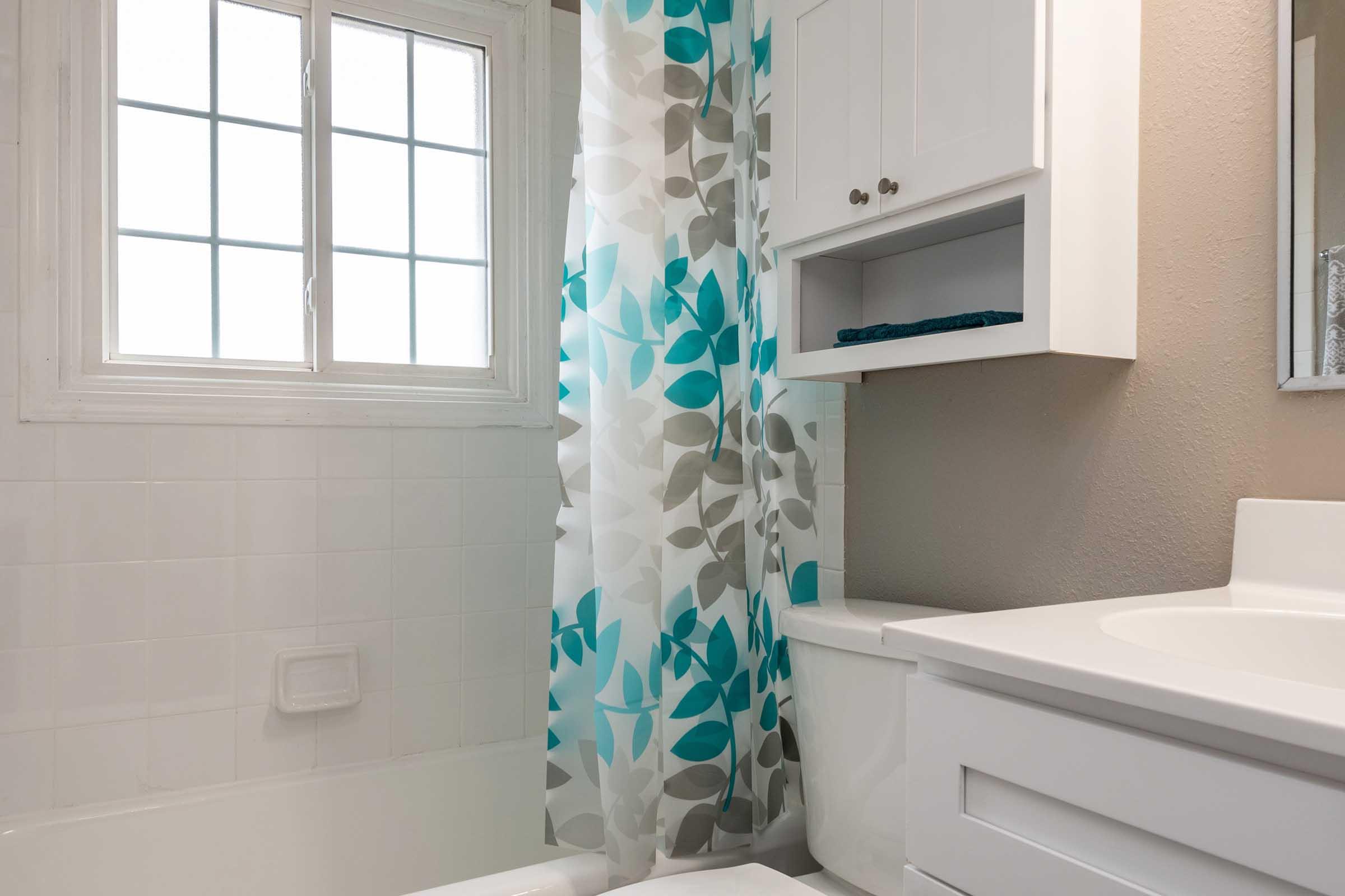 Bright and clean bathroom featuring a bathtub with a light-colored wall, a white vanity with a sink, and a window. The shower curtain displays a modern design of blue and gray leaves, adding a pop of color to the space. A small shelf can be seen above the toilet.