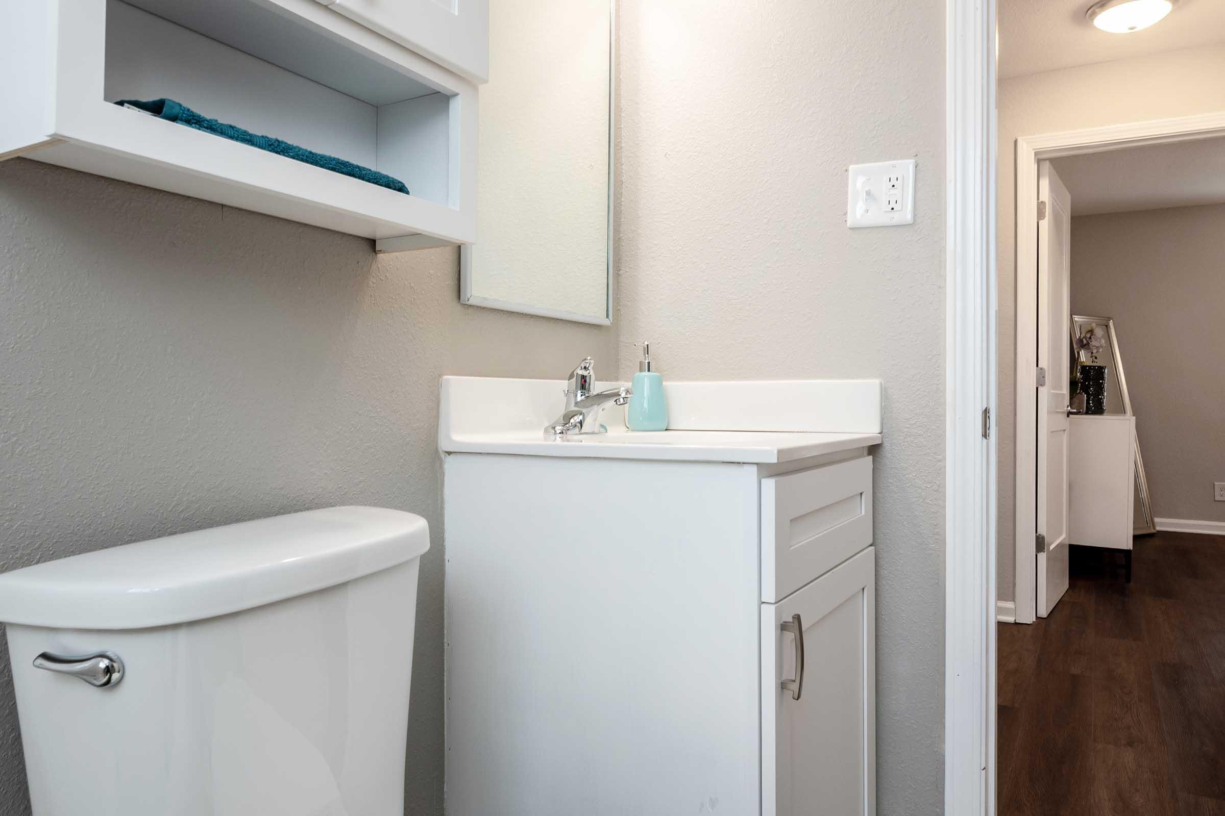 A clean, modern bathroom featuring a white vanity with a sink, a mirror above it, and a toilet. A shelf above the sink holds a rolled-up towel. The room has neutral gray walls and a wooden floor. A doorway in the background suggests connection to another room.