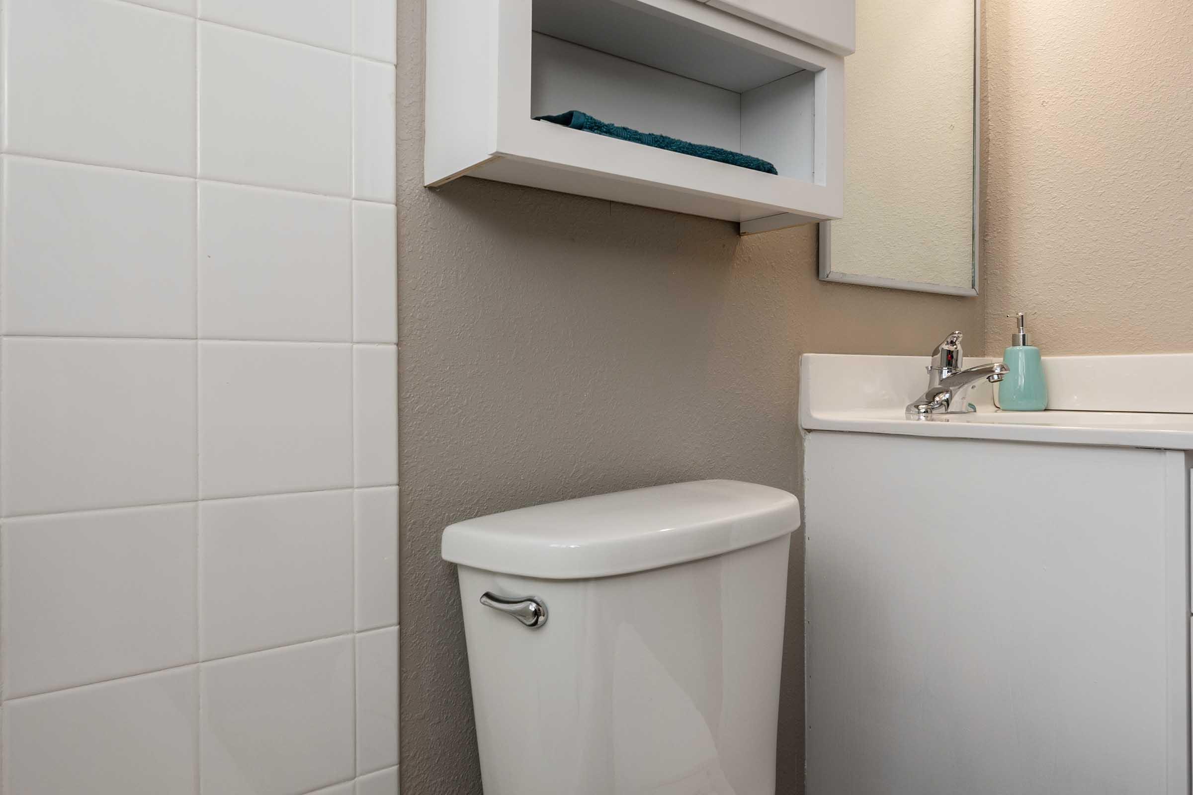 A clean bathroom featuring a white toilet, a sink with a faucet, a small shelf with a blue towel, and a mirror above the sink. The walls are painted a neutral color, and there are white tiles on one wall, creating a fresh andmodern appearance.
