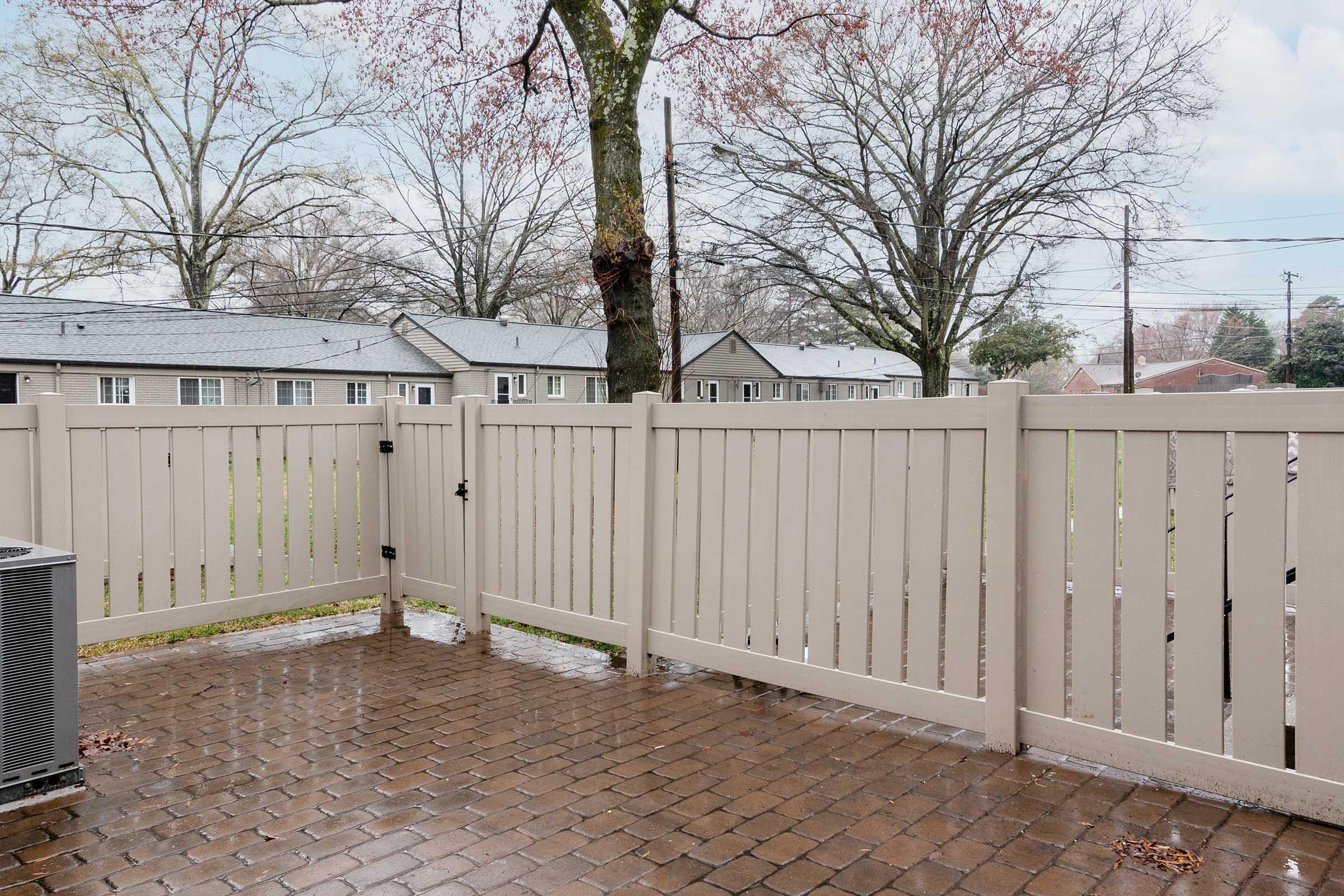 A view from a patio featuring a light-colored wooden fence surrounding a small yard. The ground is paved with brown bricks and is wet, likely from recent rain. In the background, there are several gray-roofed buildings and bare trees with dried leaves. The scene is overcast and calm.