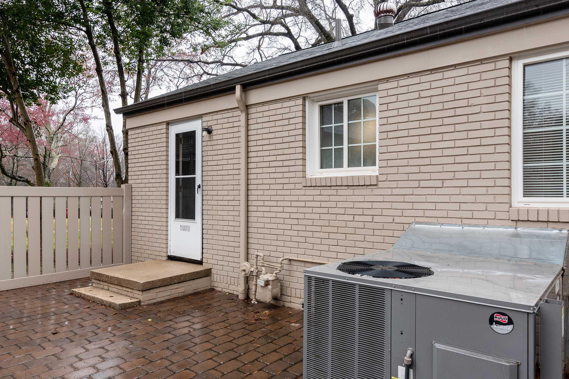 A side view of a house featuring a cream-colored brick exterior, a white door, and a window. There are steps leading up to the door, and an air conditioning unit is positioned next to the house. A wooden fence and trees are visible in the background. The ground is paved with dark brick tiles.