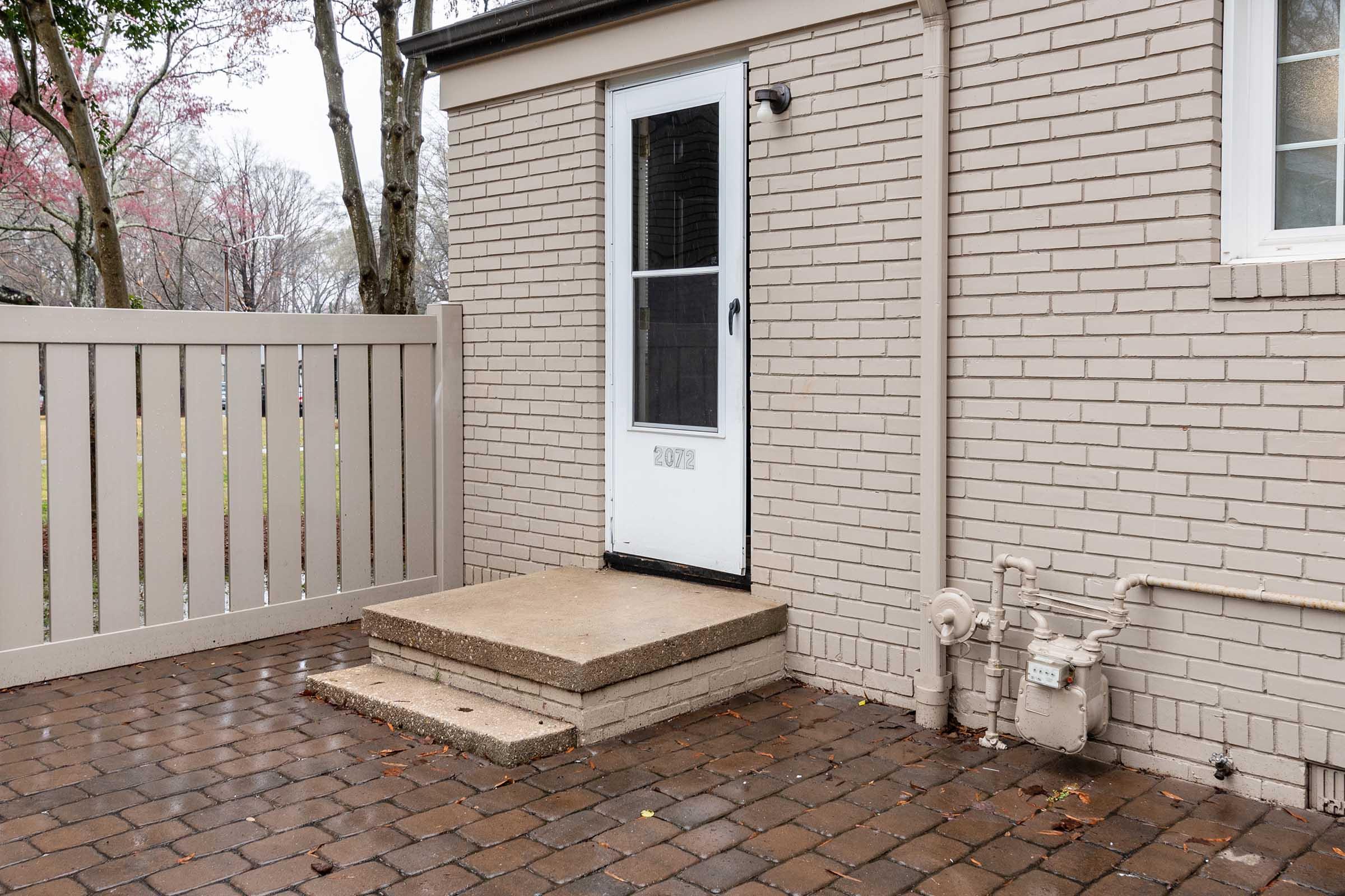 A beige brick wall with a white door and a small set of concrete steps leading to it. Beside the door, a gas meter is visible. The area features a fenced yard with a stone patio, and the ground is wet, possibly from recent rain. Trees are in the background, showing a hint of pink foliage.