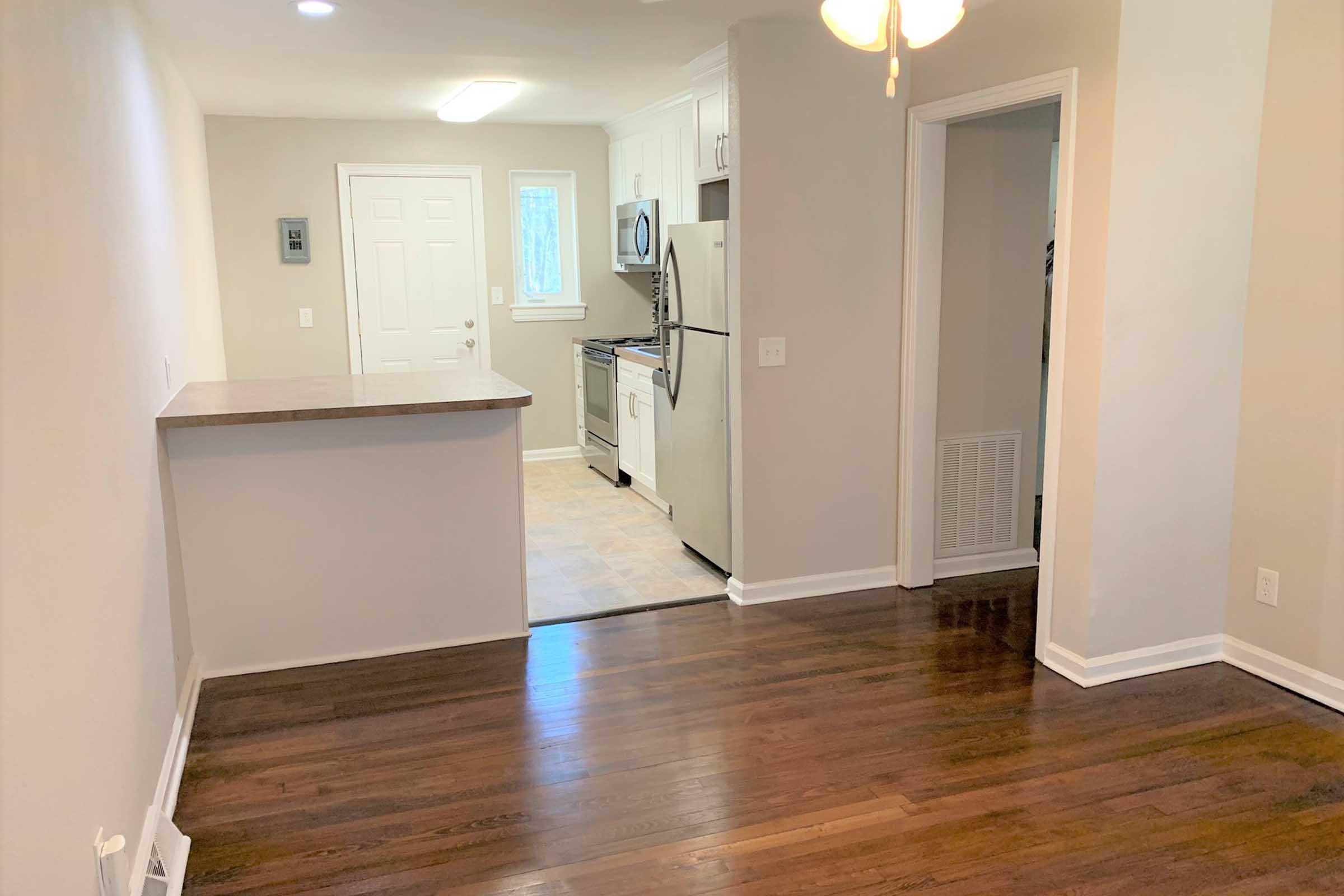 A well-lit kitchen and dining area with hardwood floors. The kitchen features white cabinets, a stainless steel refrigerator, and an adjacent doorway leading to an entrance. A small bar counter separates the kitchen from the living space. The walls are painted light gray, creating a modern and inviting atmosphere.