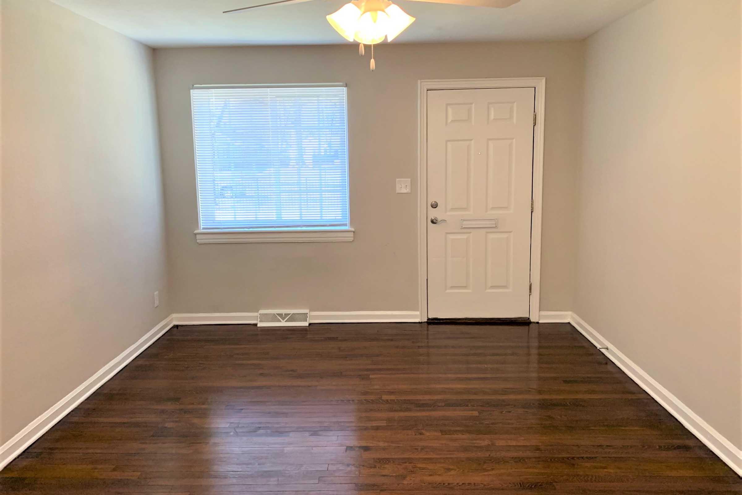 A light-filled, empty room with hardwood floors, featuring a window with blinds and a white door. The room has a ceiling fan and neutral-colored walls, creating a spacious and inviting atmosphere.