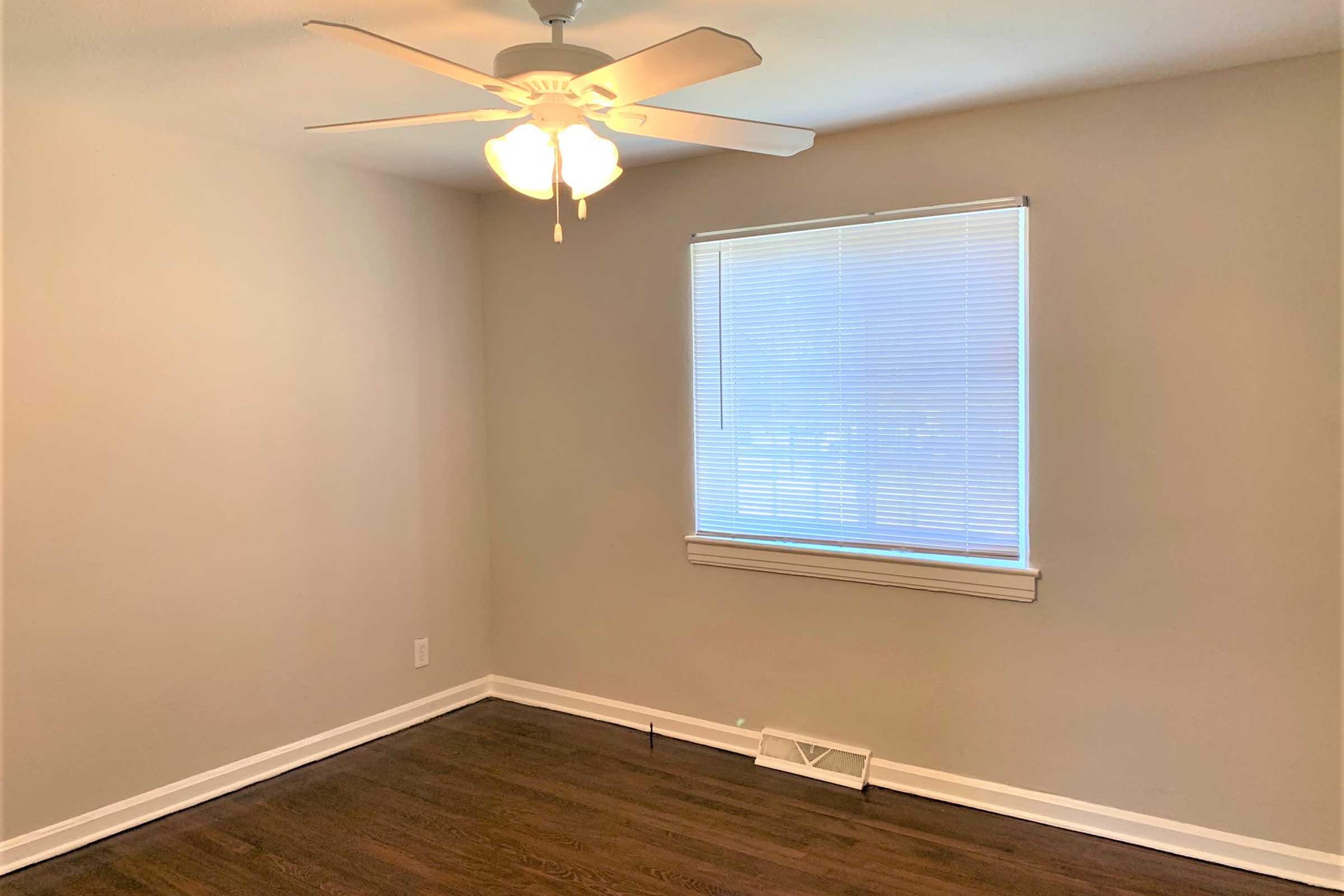 A small, empty room featuring a ceiling fan, a window with blinds, and light-colored walls. The floor is dark wood, and there is a vent on the floor. The room has a bright, airy feel due to the natural light coming from the window.