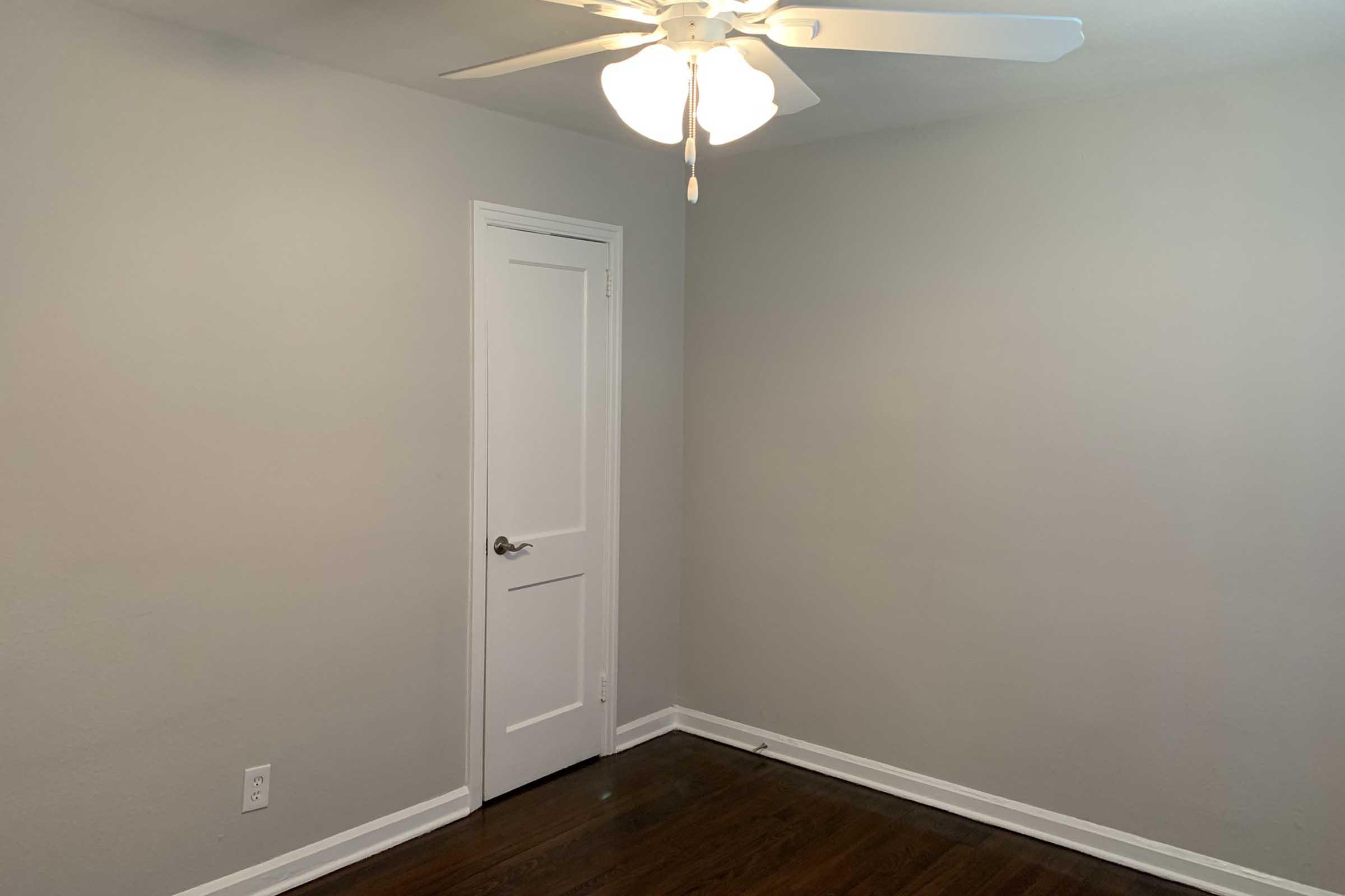 A simple, empty room with light gray walls and dark wooden flooring. A closed white door is visible on one wall, and a ceiling fan with light fixtures hangs from the ceiling. The room appears well-lit and uncluttered, ideal for various uses or decoration.