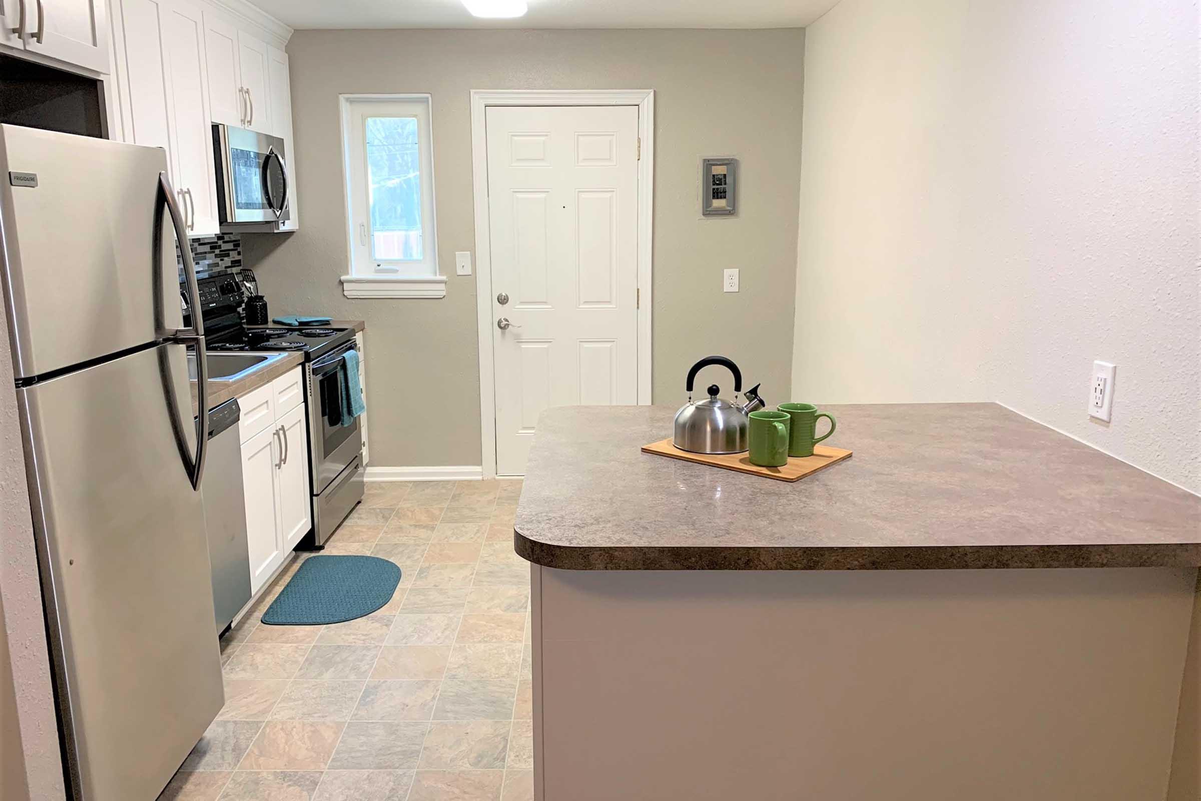 A modern kitchen featuring white cabinets, stainless steel appliances, and a light gray wall. A countertop with a kettle and two green mugs is visible in the foreground, while the entrance door and a window offer natural light. Tile flooring complements the overall clean and tidy appearance of the space.