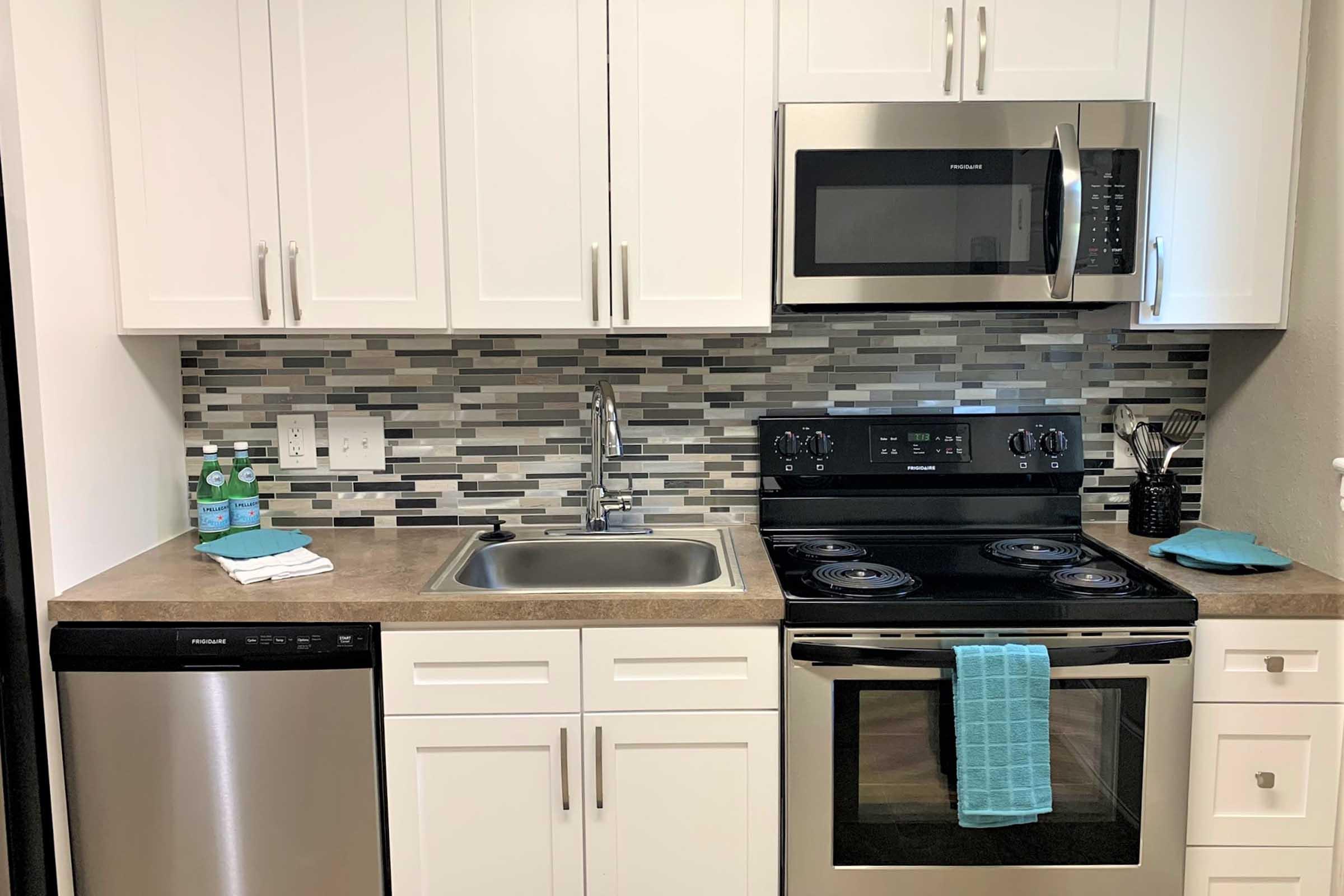 A modern kitchen featuring stainless steel appliances, including a microwave, oven, and dishwasher. The countertop is light brown with a sink and a backsplash of gray and white tiles. Blue towels are placed near the sink and on the oven, adding a pop of color to the space.