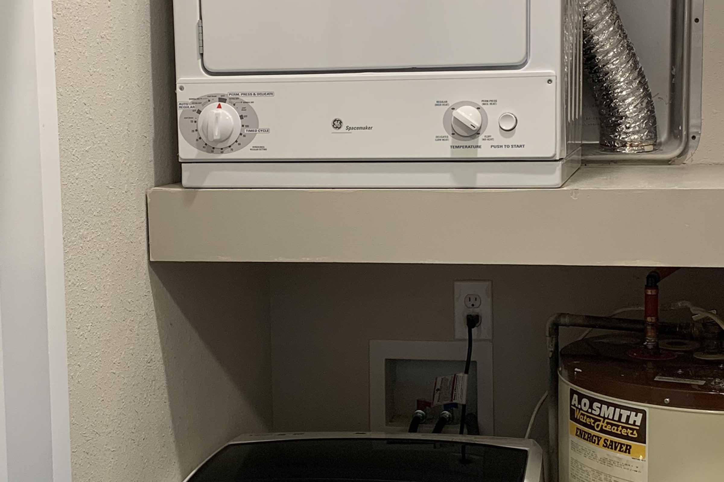 A compact laundry area featuring a stacked washer and dryer unit on a shelf. Below, there's a washing machine and a water heater with a visible label. The area is clean and organized, with plumbing and electrical connections visible on the wall.