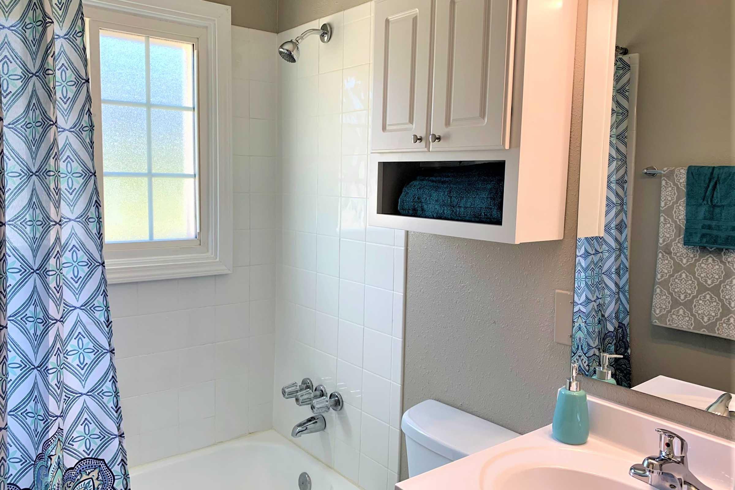 A well-lit bathroom featuring a bathtub with a shower, white tiles, and a window. There are blue patterned curtains and towels, a small cabinet with a neatly rolled towel, and a sink with a stylish soap dispenser. The overall decor is modern and welcoming.