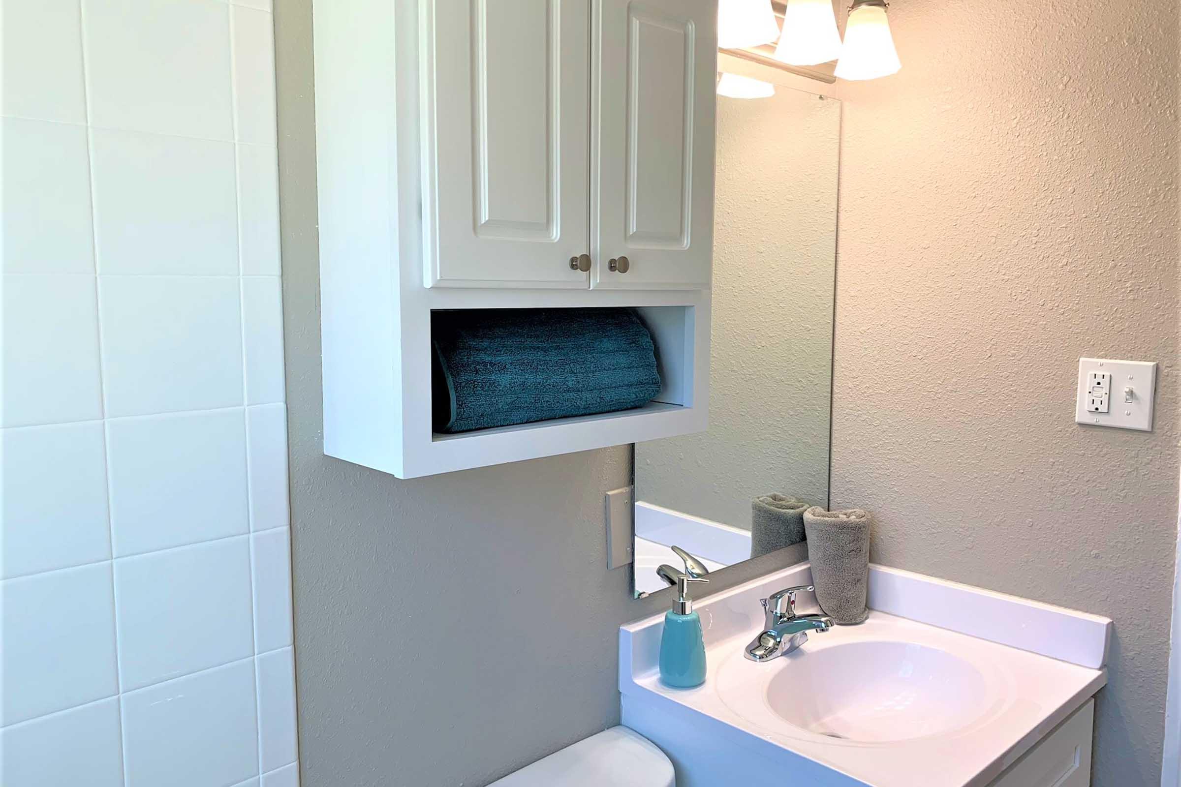 A clean and modern bathroom featuring a white vanity with a sink, a mounted cabinet containing a rolled blue towel, and a mirror above. The walls are painted a soft beige, and the bathroom has a neutral color scheme with a hint of blue from the towel and a small decorative bottle near the sink.
