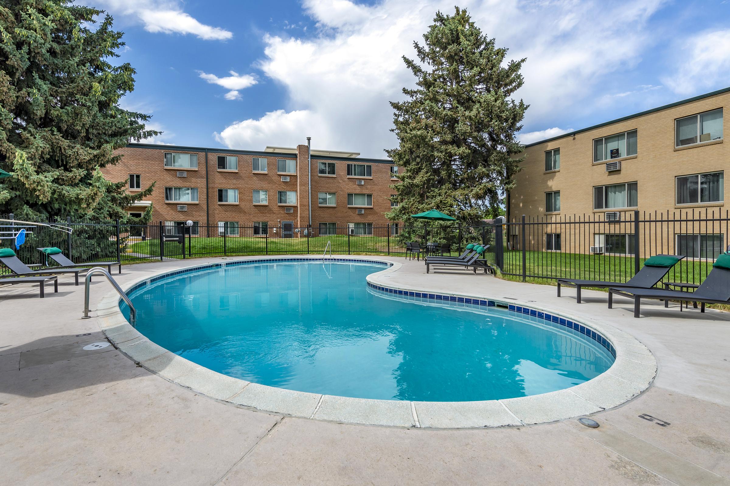A curved swimming pool surrounded by lounge chairs and umbrellas, set against a backdrop of two brick apartment buildings and greenery. The sky is partly cloudy, creating a bright and inviting atmosphere.
