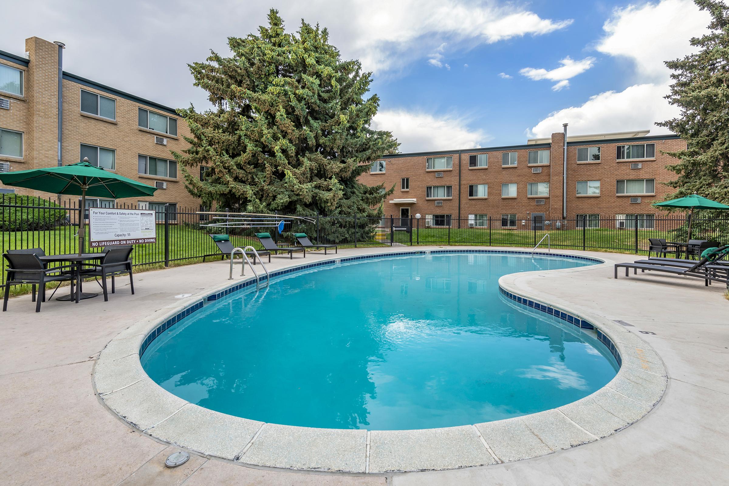 A tranquil outdoor swimming pool surrounded by lounge chairs and umbrellas, with a backdrop of a brick apartment building and green trees. The water is clear, reflecting the blue sky and clouds, creating a relaxing atmosphere for residents and visitors.