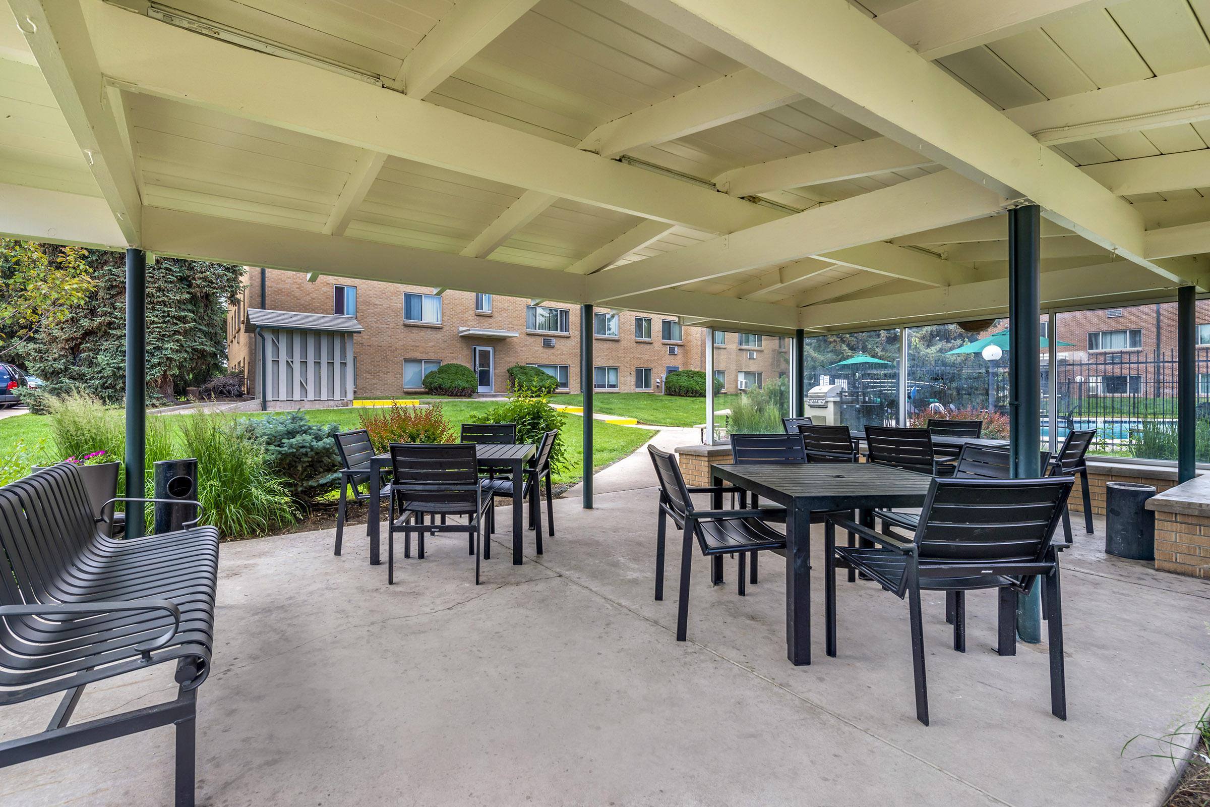 A covered outdoor seating area with several black tables and chairs, surrounded by greenery. In the background, a brick building and a pool area are visible. The space is well-lit and provides a relaxing environment for gatherings or meals.