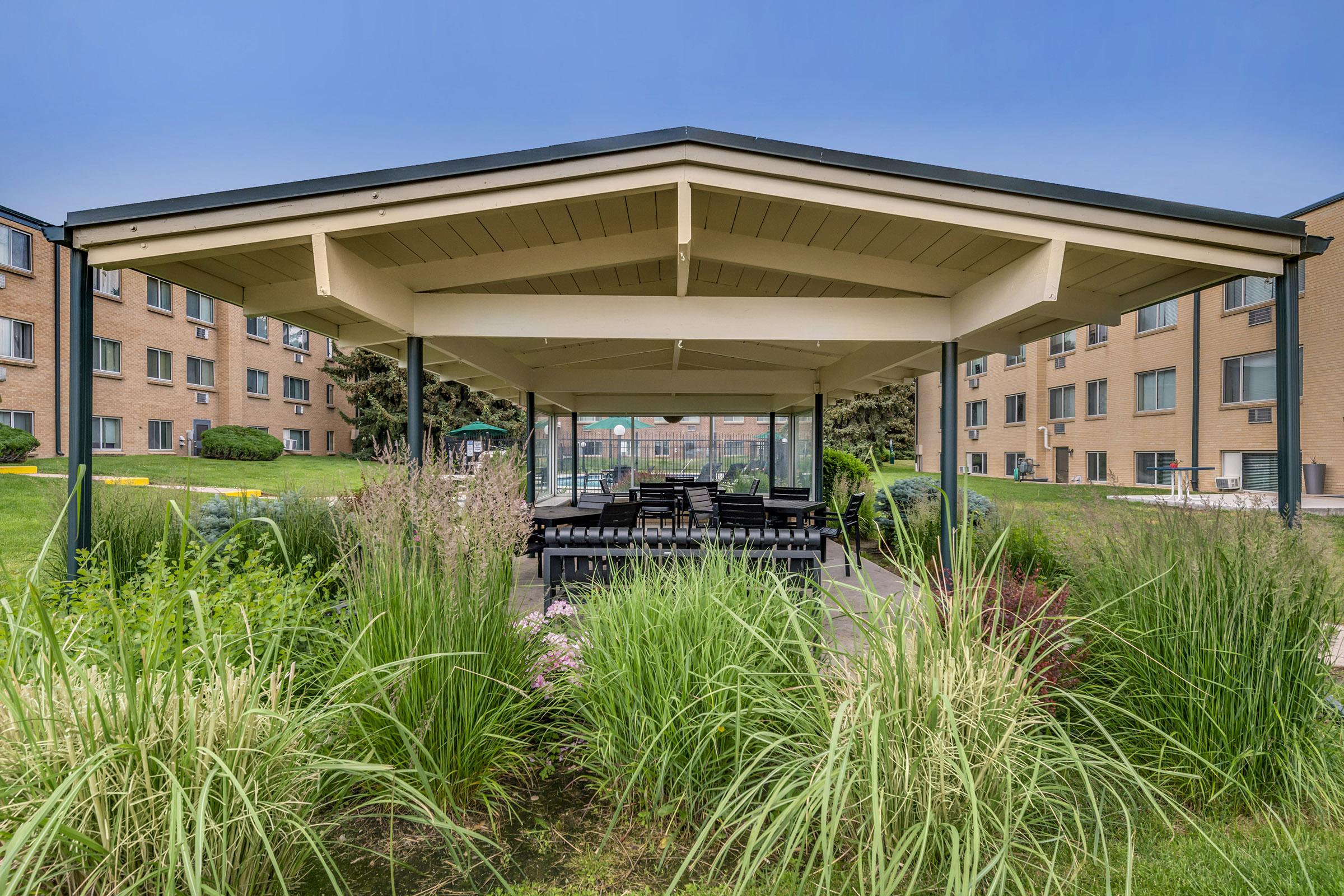 A well-structured pavilion with a peaked roof is surrounded by lush greenery and colorful plants. Inside, there are several black picnic tables arranged for gatherings. In the background, a brick building and a clear sky can be seen, creating a serene outdoor space.