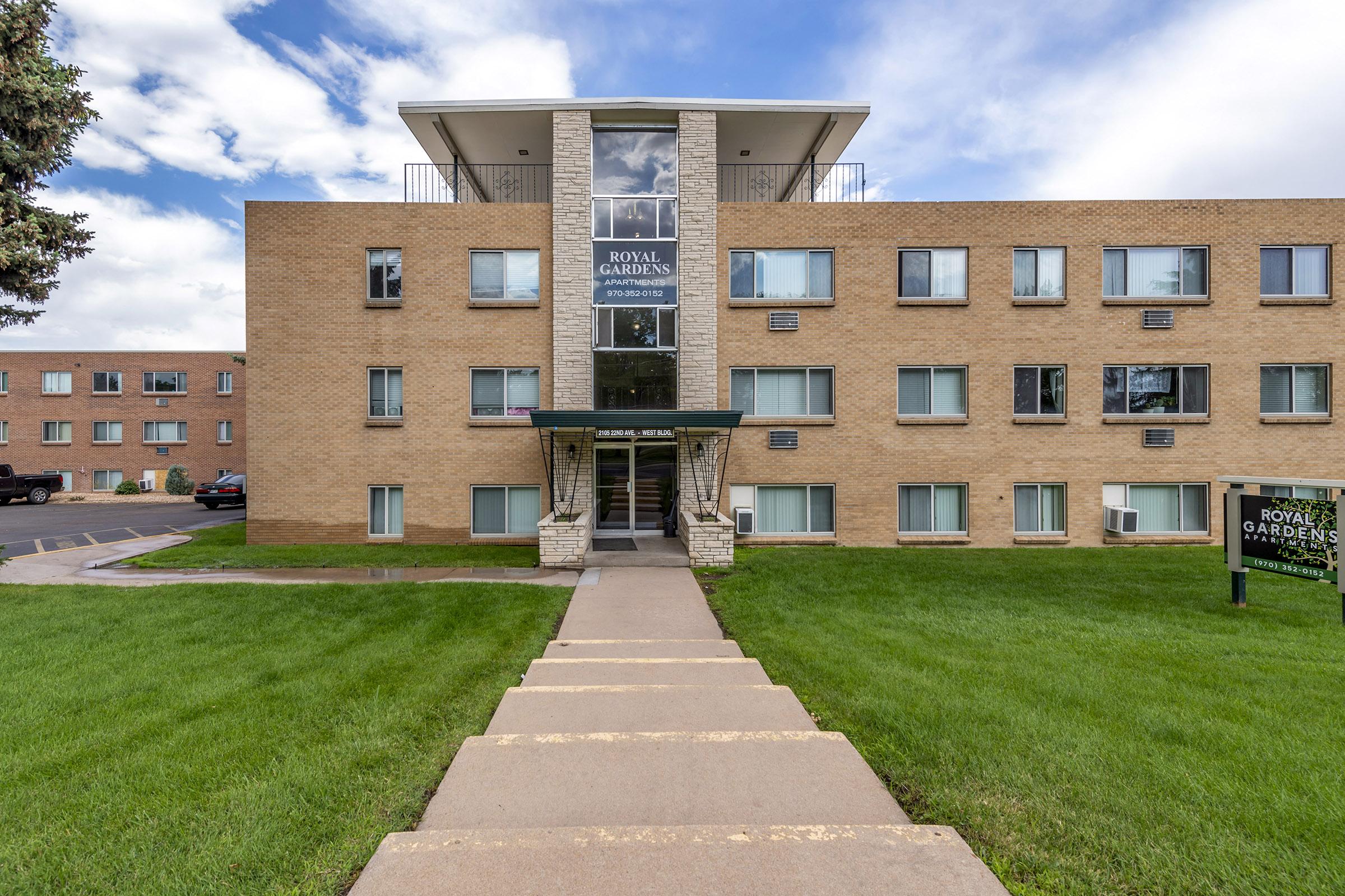 Exterior view of a multi-story brick apartment building named "Royal Gardens." The entrance features a small awning and a pathway leading to it. Surrounding the building is a grassy area with trees, under a partly cloudy sky.