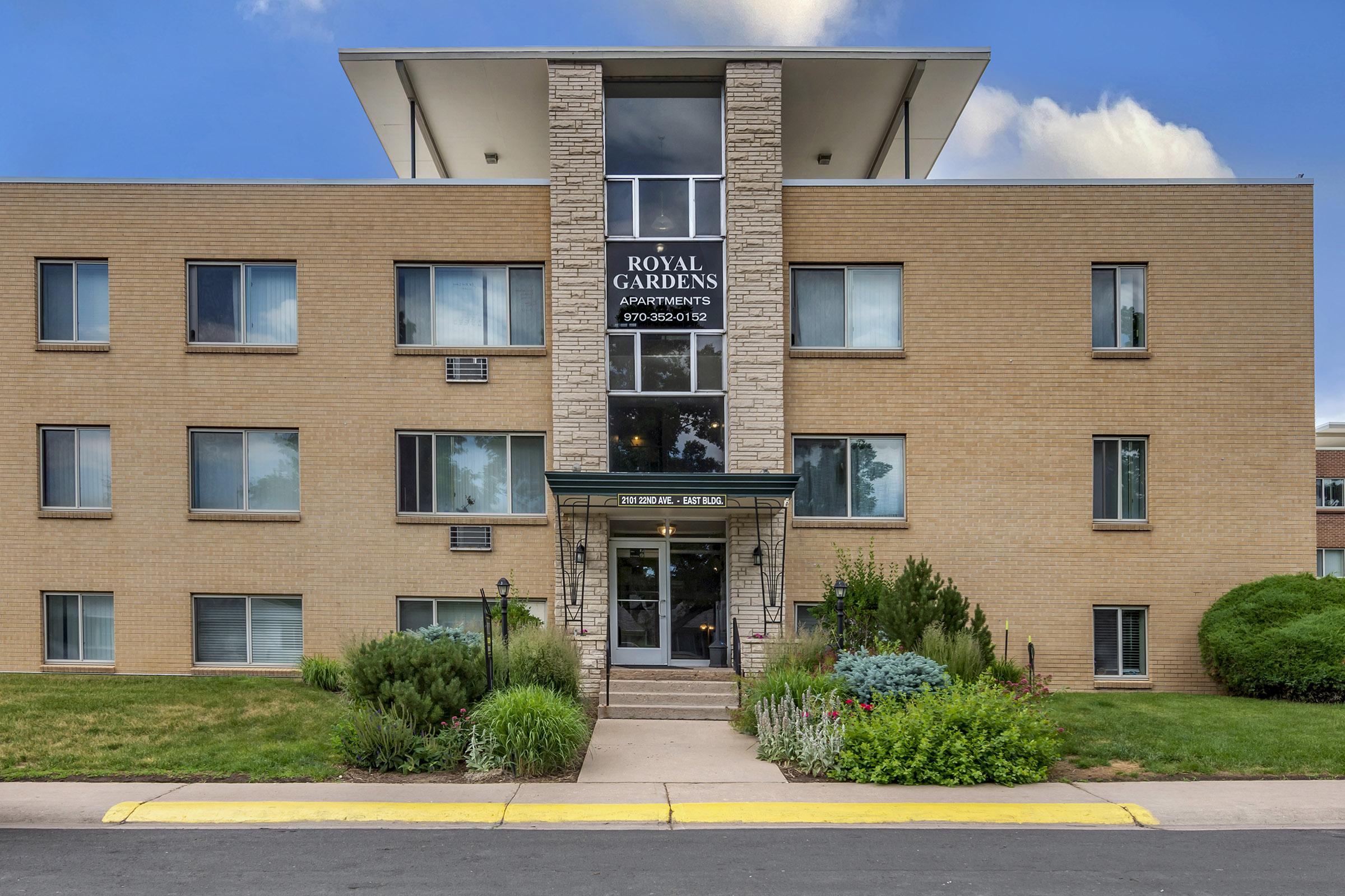 Exterior view of Royal Gardens Apartments, featuring a three-story brick building with large windows and a welcoming entrance. Lush greenery and flower gardens are visible in the foreground, with a clear blue sky above. The building has a modern design, making it an attractive residential option.