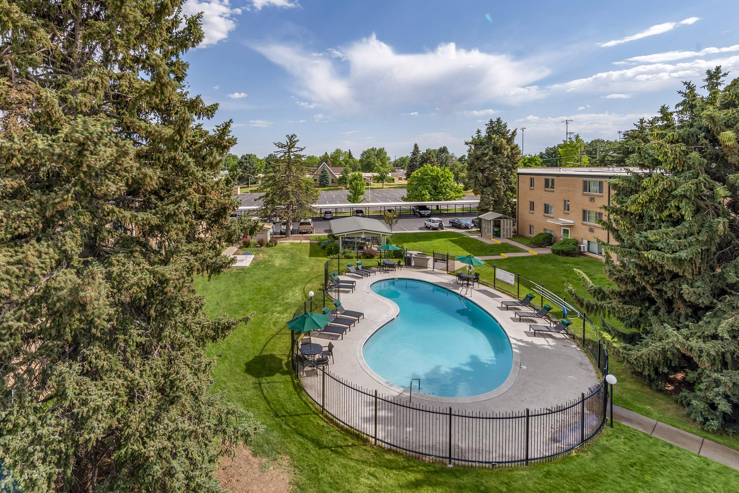 Aerial view of a landscaped area featuring a rectangular swimming pool surrounded by green grass and trees. There are lounge chairs and umbrellas around the pool, with nearby apartment buildings. The sky is mostly clear with a few clouds.