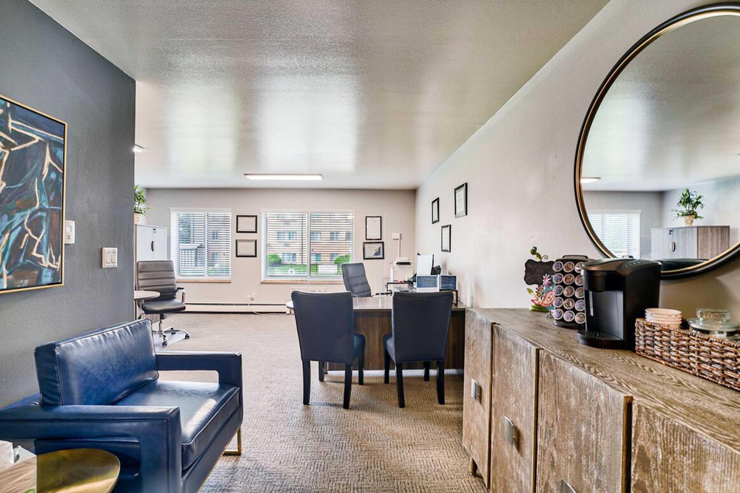 A modern office space featuring a brown wooden desk with a computer, two black chairs, and a blue leather chair. There is a circular mirror on the wall, framed art, and a coffee station. The room has large windows allowing natural light, and greenery adds a touch of decor.