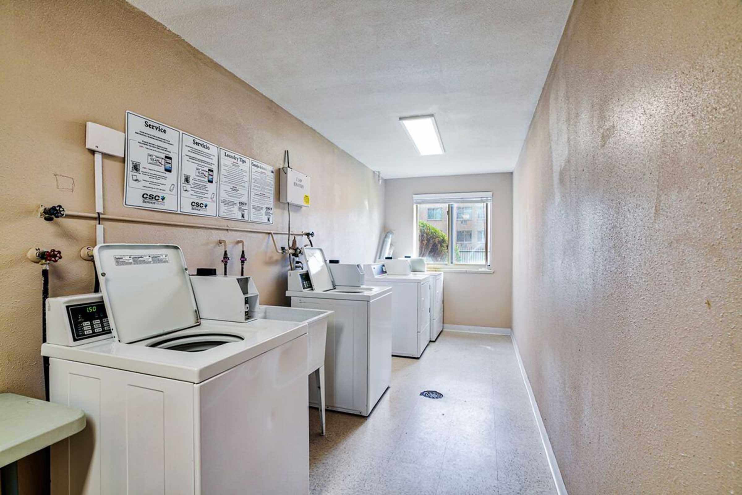 A clean and well-lit laundry room featuring several white washing machines and a dryer along one side. The walls are a neutral beige color, and there are informational posters on the wall about laundry services. A window lets in natural light, providing a welcoming atmosphere.