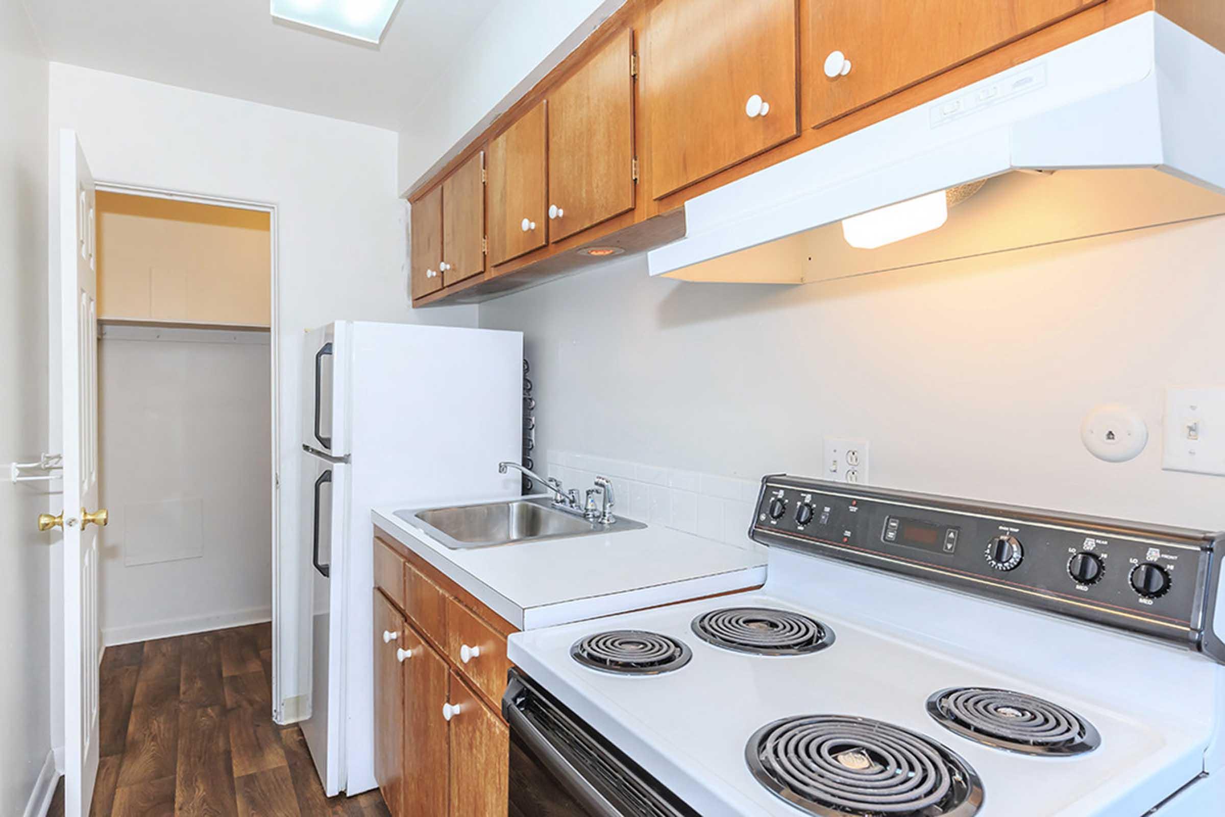 A compact kitchen featuring wooden cabinets, a white sink, a stovetop with four burners, and an oven. The kitchen includes a refrigerator on the left and a light-colored wall, with a door leading to a small storage area in the background. The flooring is a light wood laminate.
