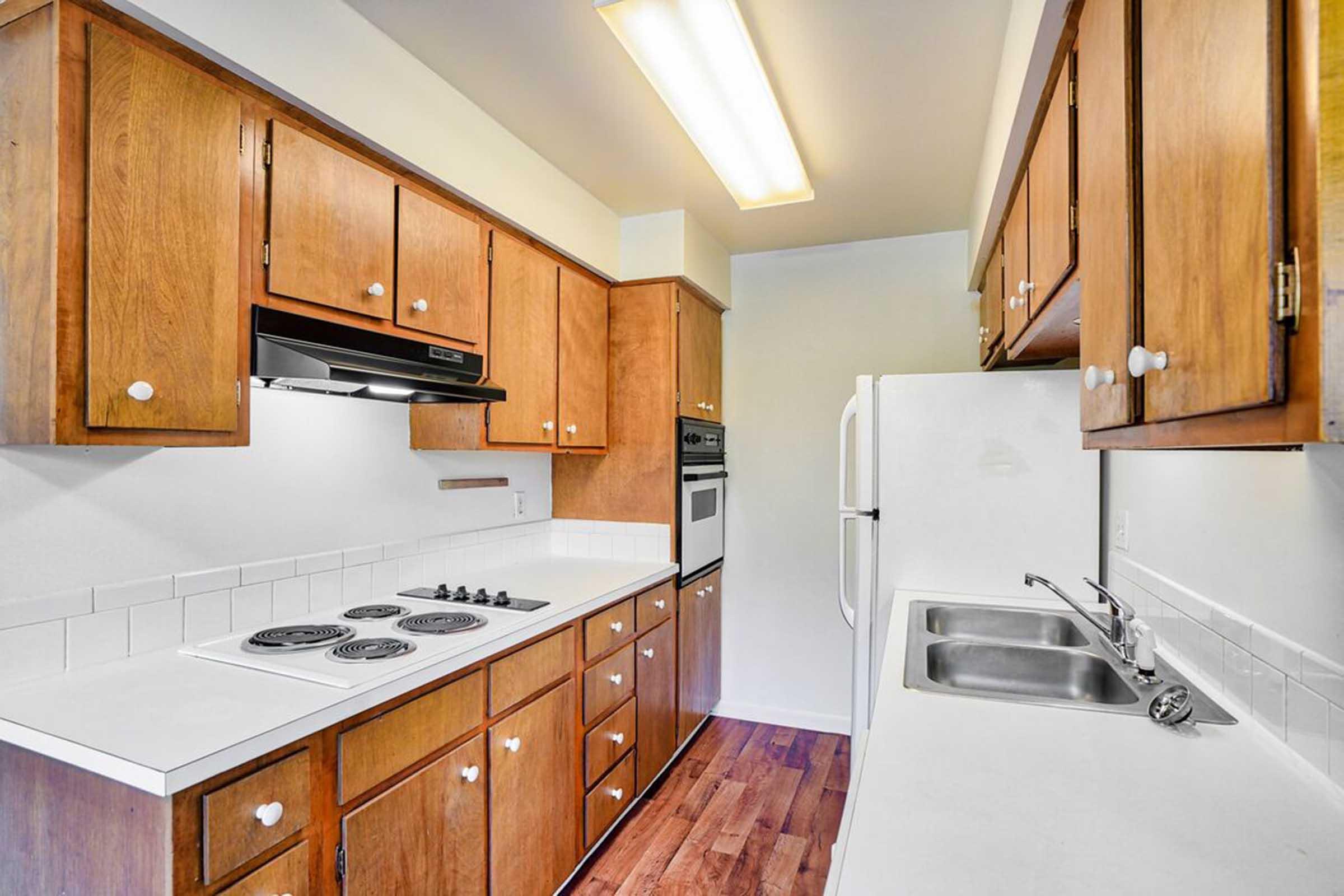 A modern kitchen featuring wooden cabinets, a white countertop, a double sink, a stove with four burners, an oven built into the cabinetry, and a refrigerator. The space is well-lit with overhead lighting and has a clean and organized appearance.