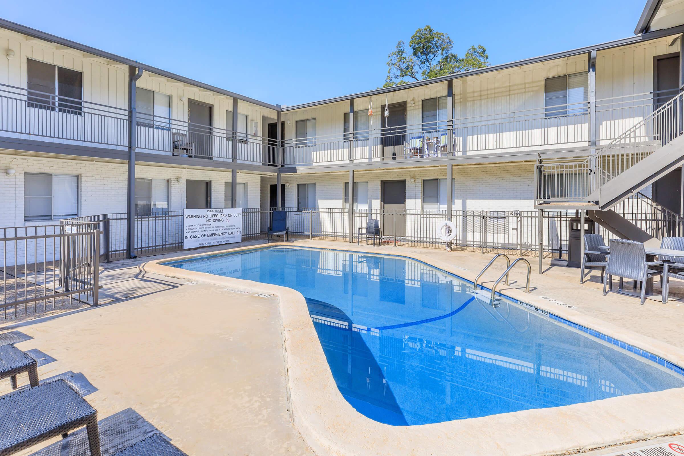 A view of a hotel courtyard featuring a swimming pool surrounded by two-story accommodation buildings. The pool is clear and inviting, with a shallow edge and a ladder. Several outdoor chairs and tables are arranged near the pool, and there are signs and balconies visible in the background. Bright blue sky overhead.