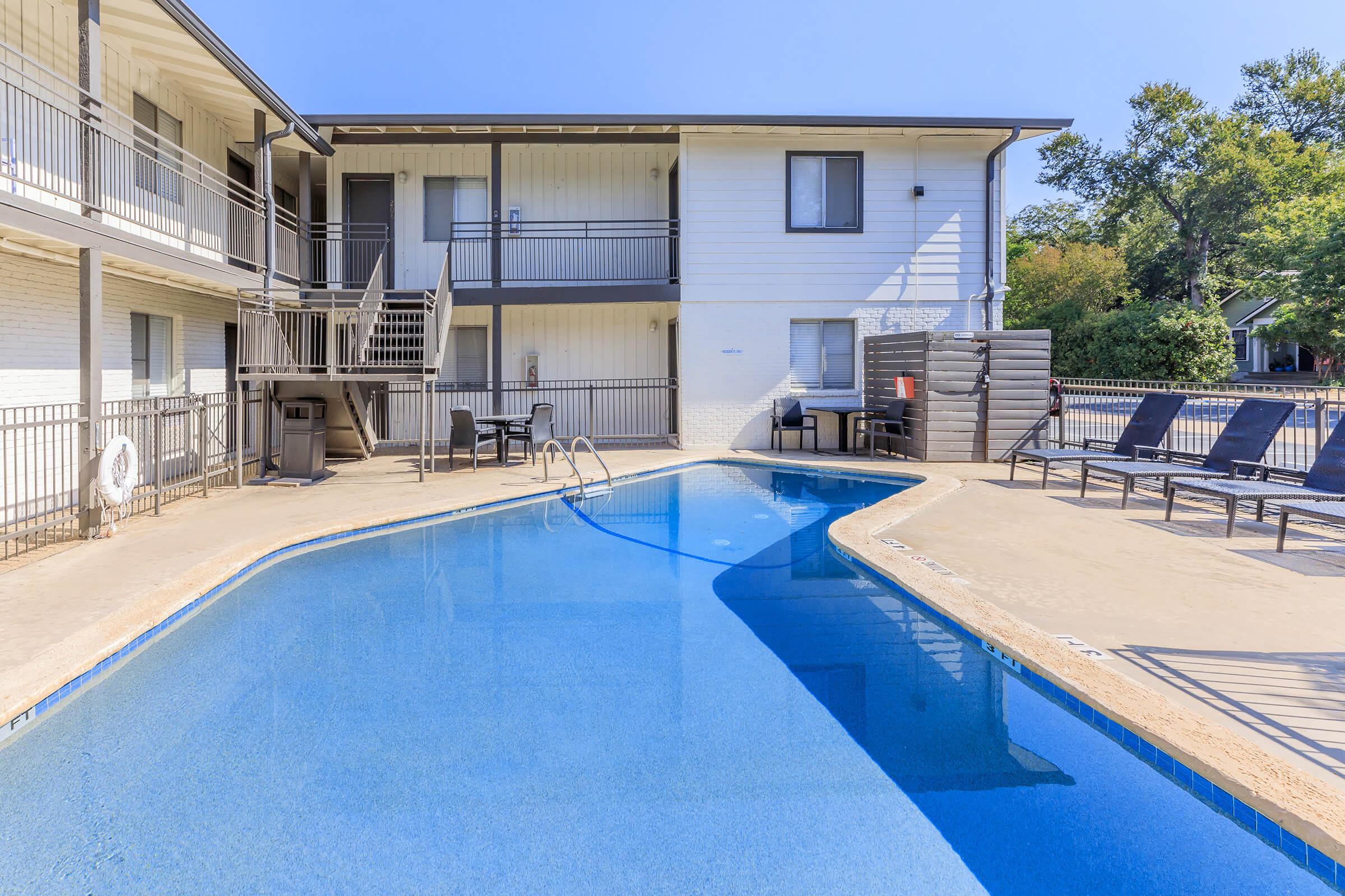 A view of an outdoor swimming pool surrounded by lounge chairs, with a two-story apartment building in the background. The building features balconies and stairs leading to the upper level, under a clear blue sky. The pool reflects the sunlight, creating a relaxing atmosphere.