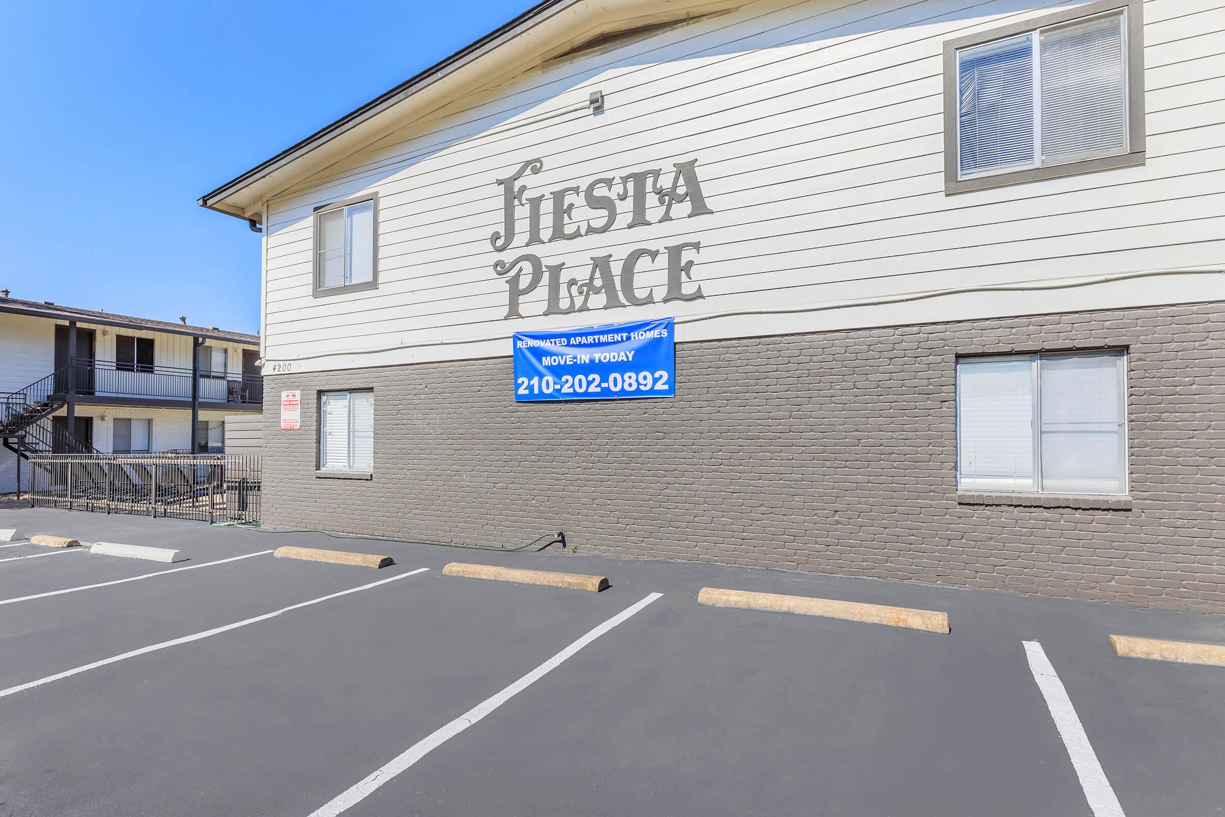 Building exterior with the sign "Fiesta Place" prominently displayed above a beige wall. A blue banner advertising a phone number for leasing information hangs below the sign. The parking area in front is paved and marked with parking spaces, with a clear blue sky visible above.