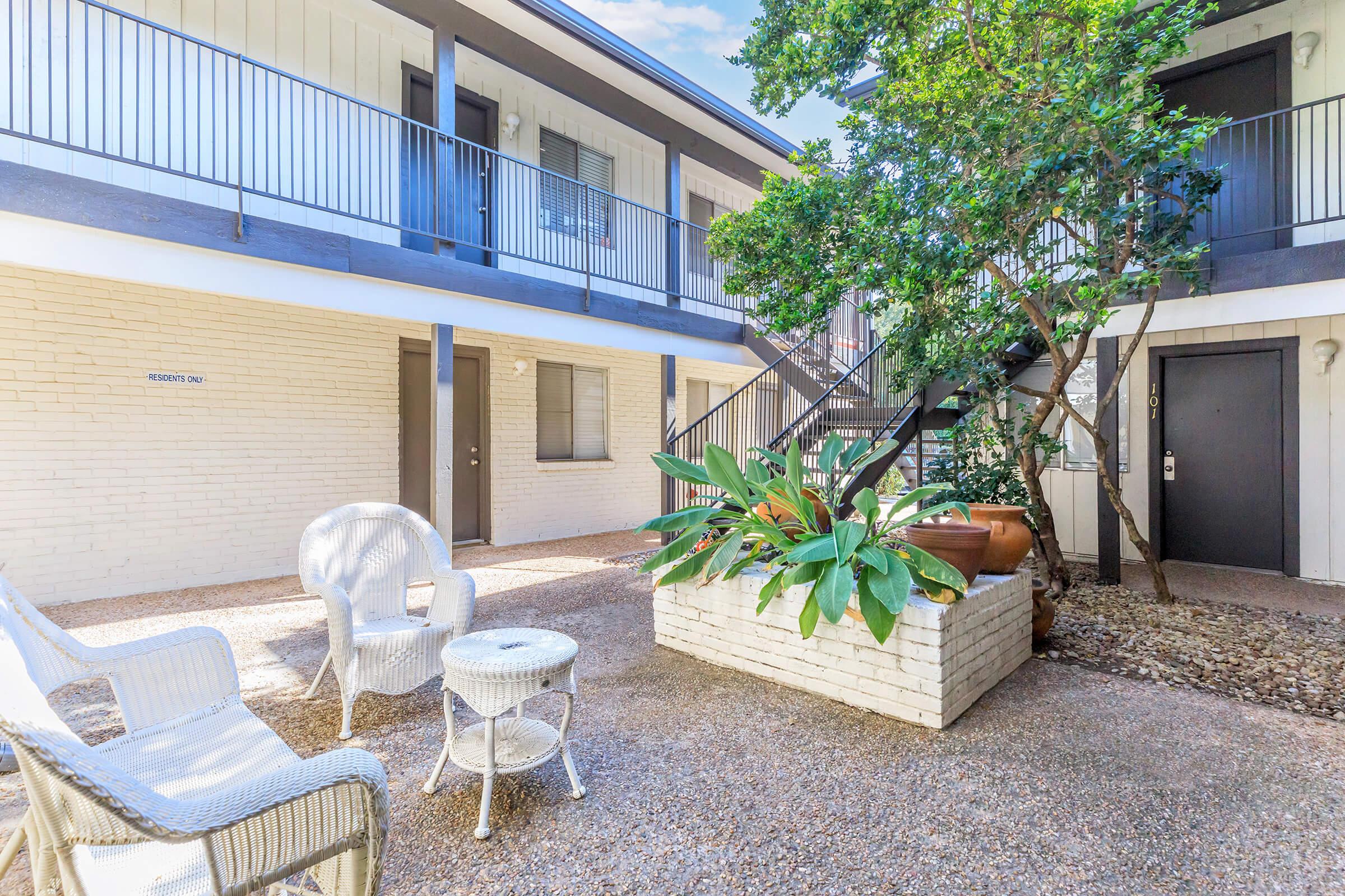 A tranquil courtyard featuring white wicker furniture, potted plants, and a small climbing staircase. The space is surrounded by two-story apartments with balconies, creating a serene outdoor atmosphere. Gravel pathways and greenery enhance the inviting ambiance.
