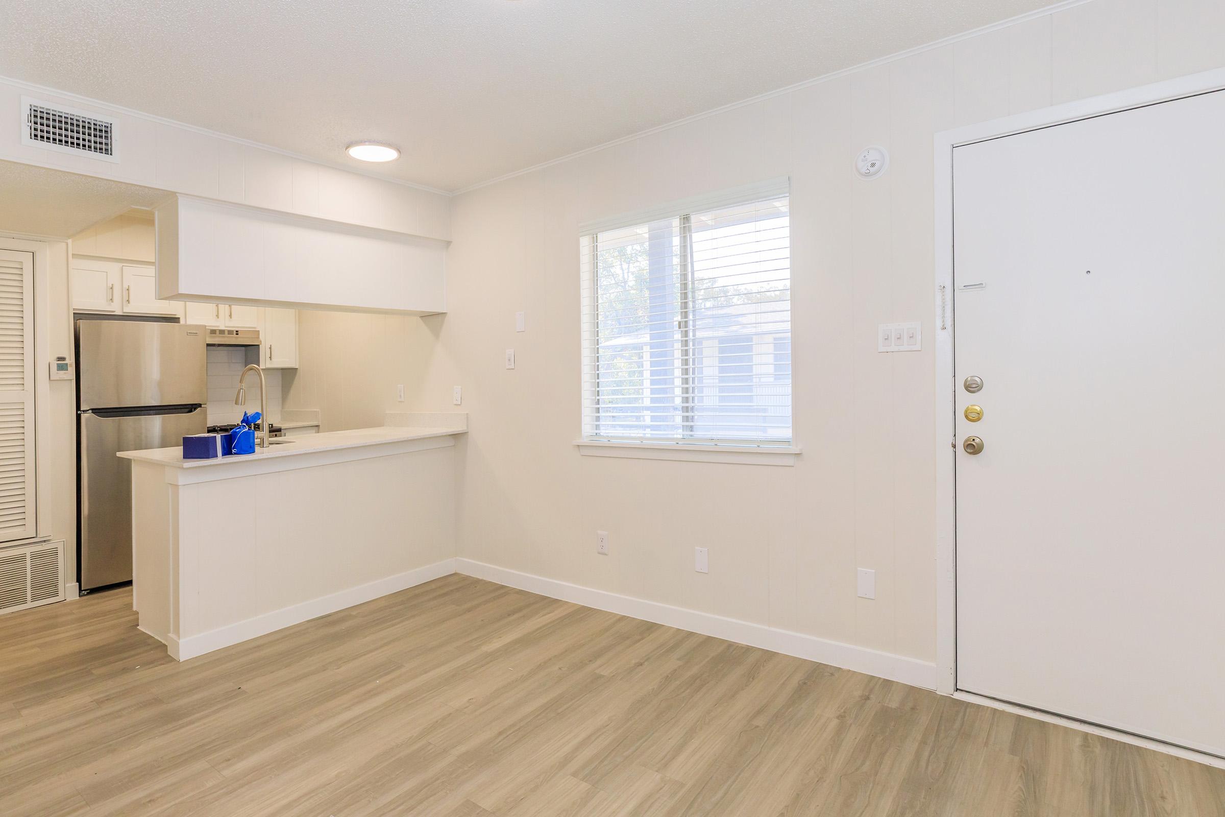 A bright, modern living space featuring light-colored walls and wood-style flooring. A kitchenette with white cabinetry is visible on the left, alongside a stainless steel refrigerator. A window allows natural light to fill the room. The entrance door on the right is plain and white.