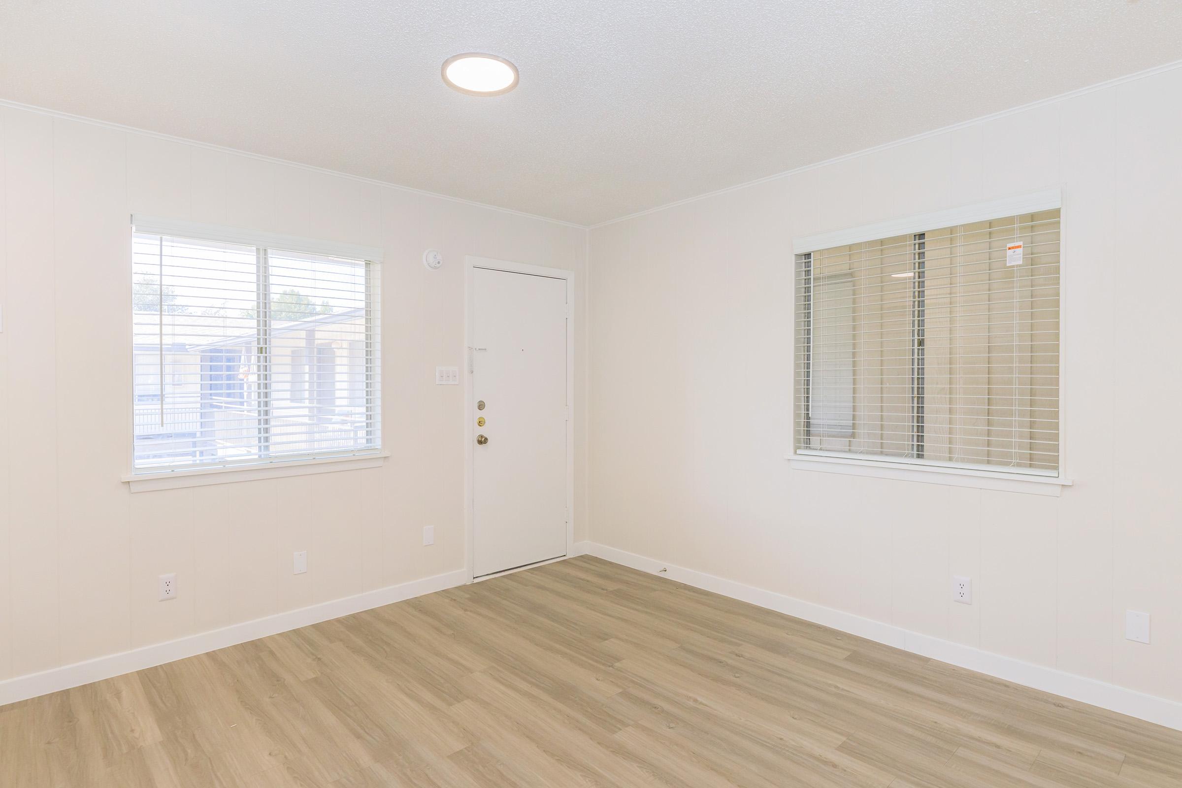 A light and airy room with a neutral color palette, featuring two windows with blinds. The walls are painted white, and the floor is made of light-colored wood. A ceiling light fixture is visible, and the door is located on the left side of the image. The overall atmosphere is clean and inviting, suitable for various uses.