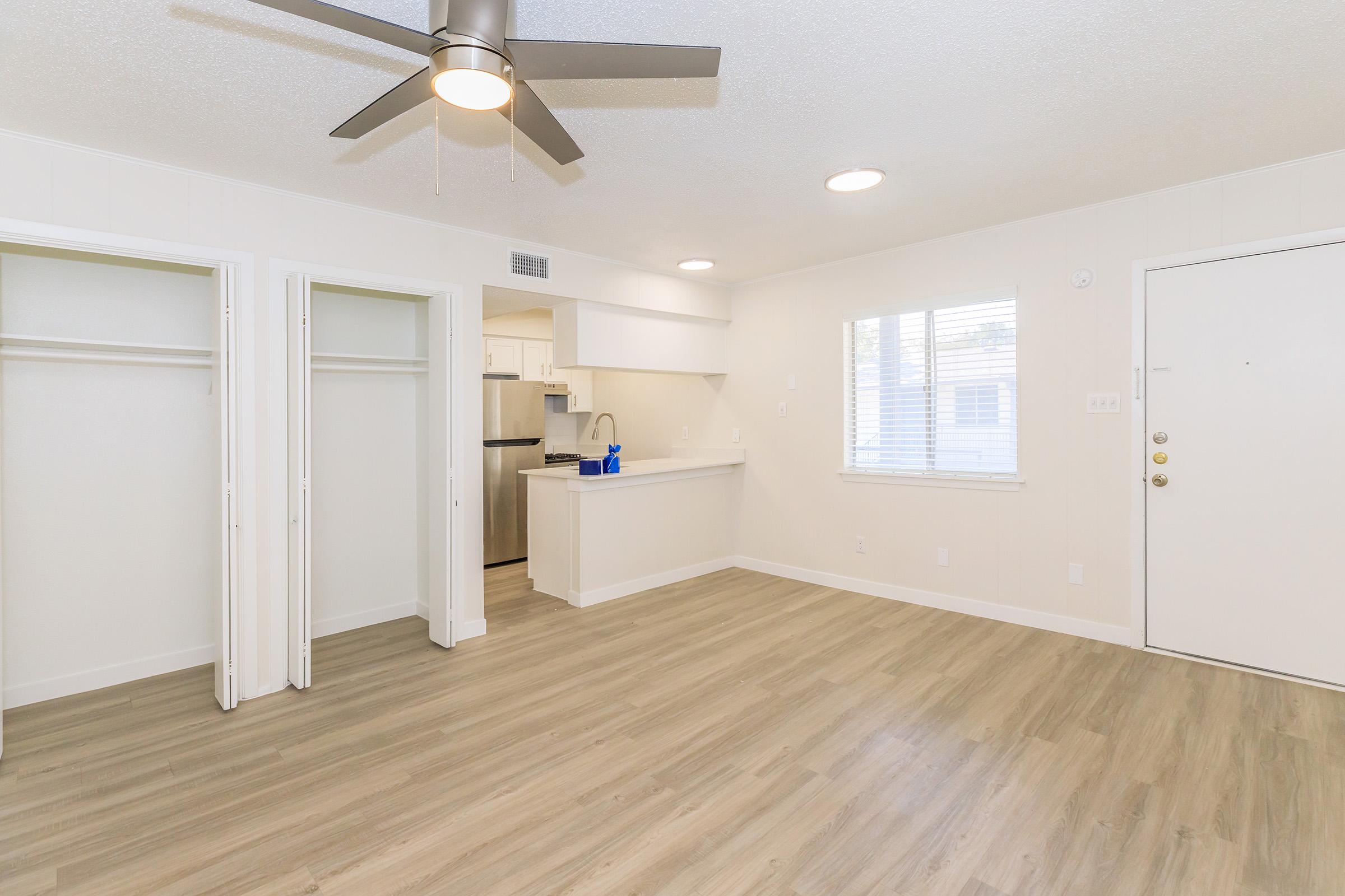 A bright, modern living space featuring light-colored walls, laminate flooring, and a ceiling fan. There are two open closets on the left, a window allowing natural light, and a kitchen area visible in the background. The entrance door is on the right, complementing a spacious and inviting atmosphere.