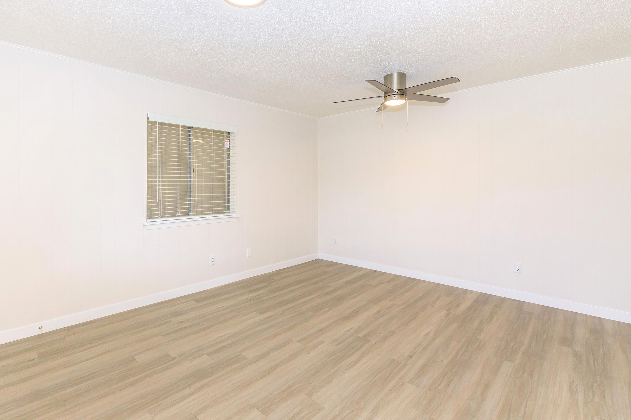 A spacious, empty room with light-colored walls and a ceiling fan. There is a window with blinds on the left side, allowing natural light to enter. The floor features light wood-like laminate, adding to the bright and airy feel of the space.