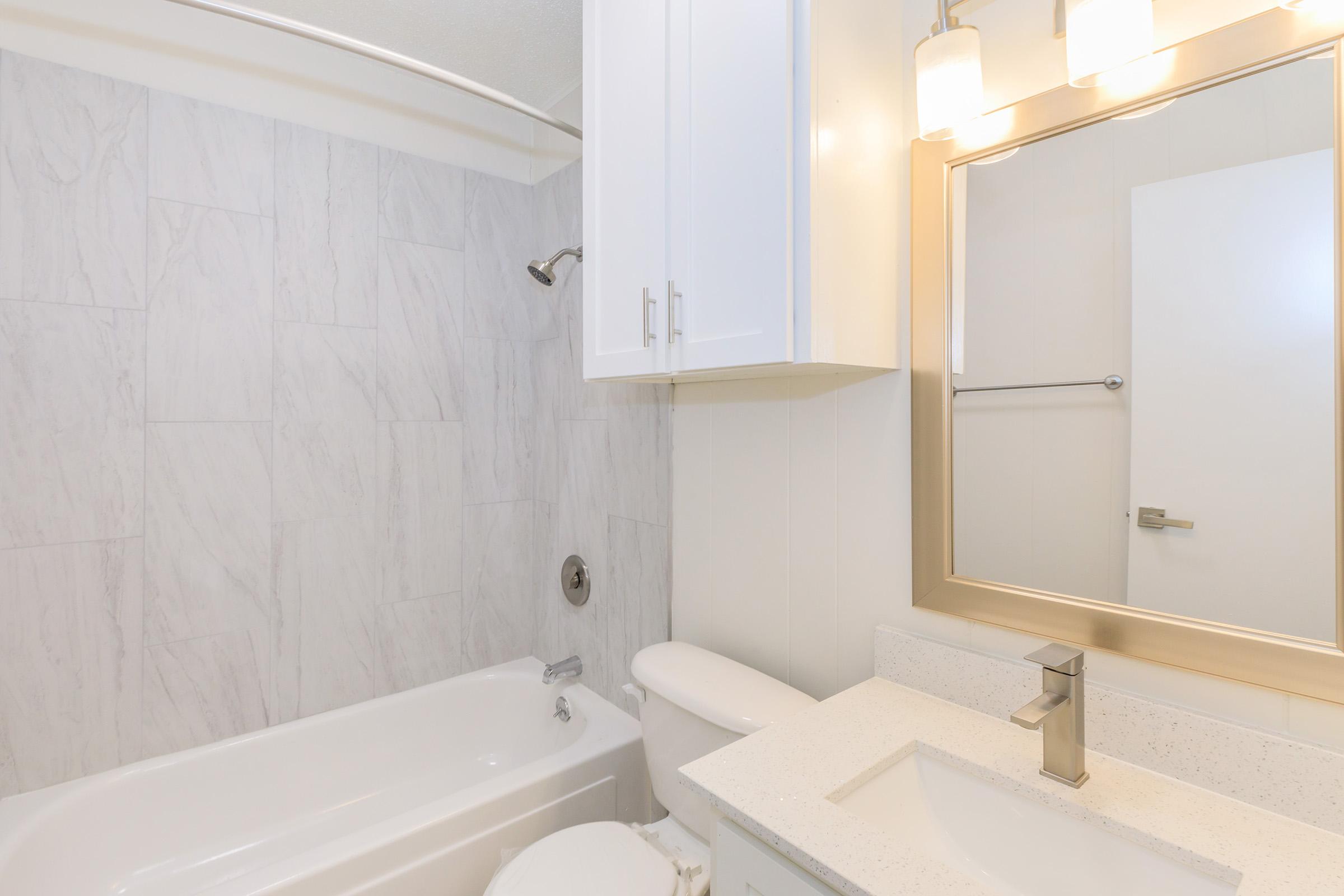A modern bathroom featuring a white bathtub, a sleek countertop with a sink, and a decorative mirror. The walls are tiled in light gray, and there are white cabinets for storage. The lighting fixtures above the mirror provide a warm glow, creating a bright and inviting space.