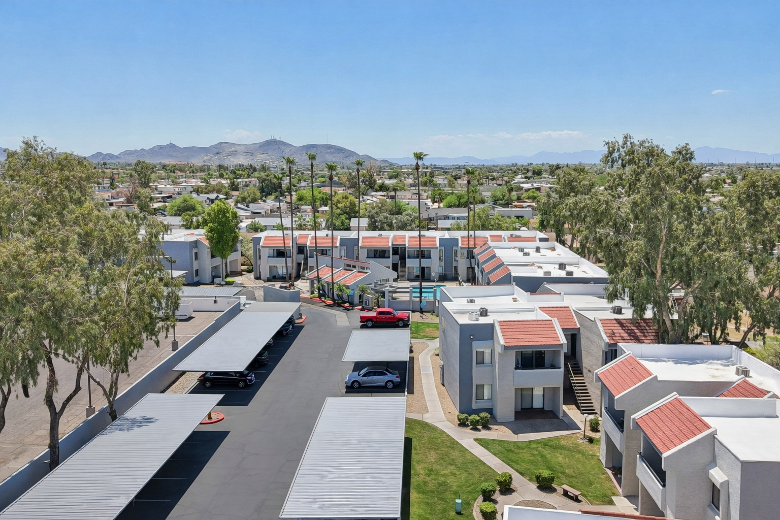Aerial view of an apartment complex with flat-roofed buildings, a central pool area, and several palm trees. There are parked cars under shaded carports and mountains visible in the background under a clear blue sky. The scene depicts a sunny residential environment.