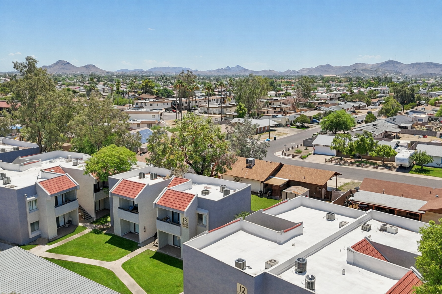 Aerial view of a residential neighborhood featuring multiple apartment buildings with flat rooftops and red tile accents. Green lawns and trees are visible between the buildings. In the background, a range of low mountains can be seen under a clear blue sky. The scene depicts a suburban setting with diverse housing styles.
